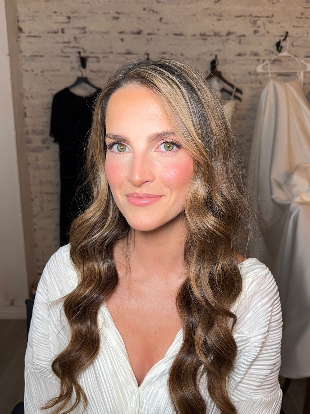 Bridal beauty portrait featuring soft waves and natural glam makeup against brick wall.
