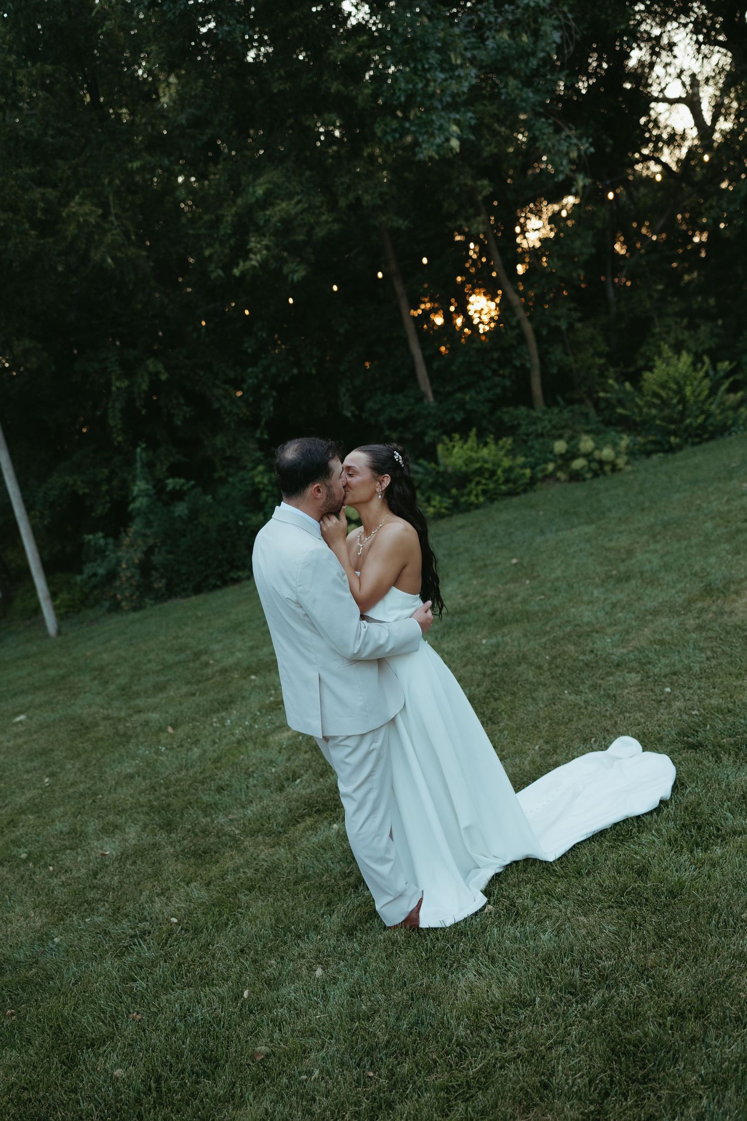 Romantic wedding couple embraces on grassy hillside at dusk.