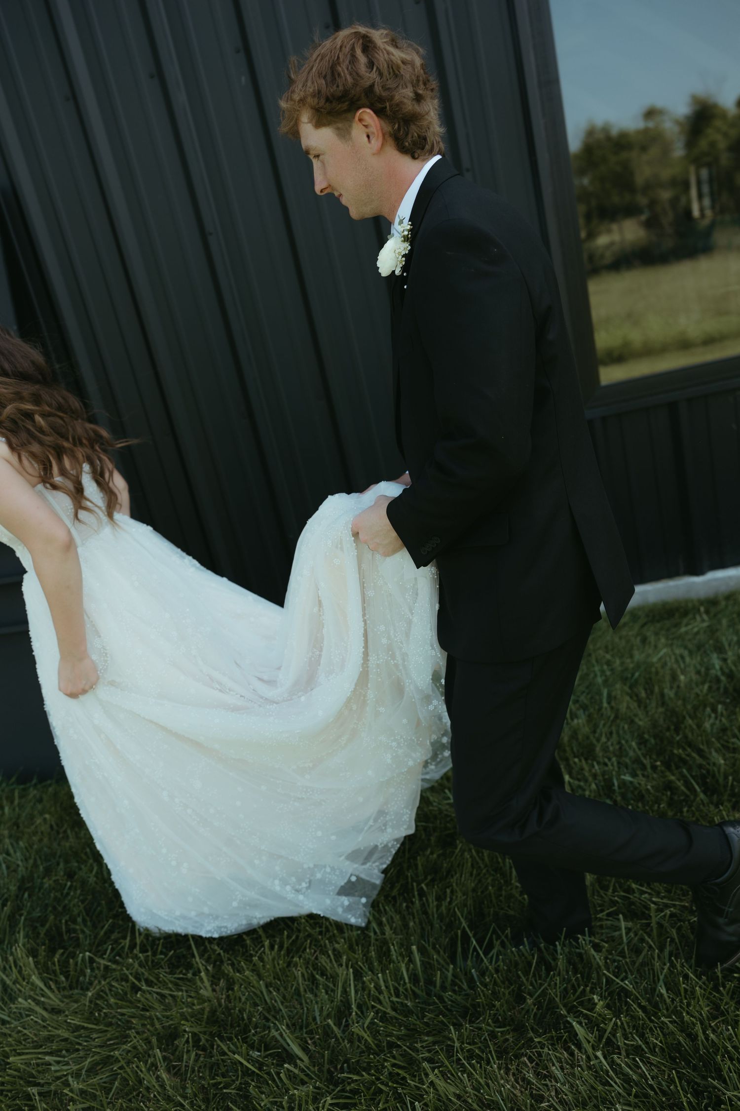 White wedding dress flows gracefully against dark exterior wall and green grass.