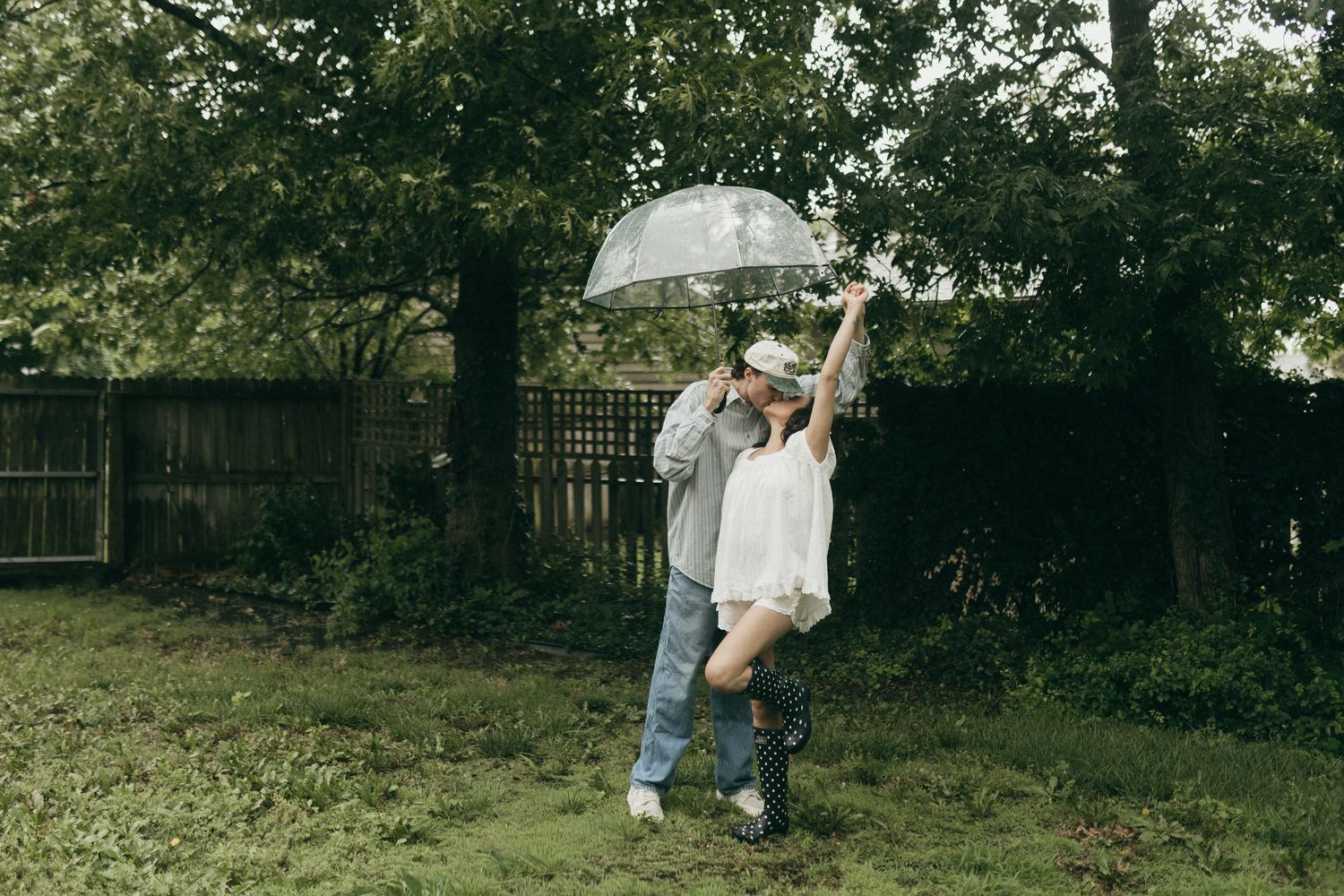Couple sharing a romantic moment under a clear umbrella in a lush green backyard on a rainy day.