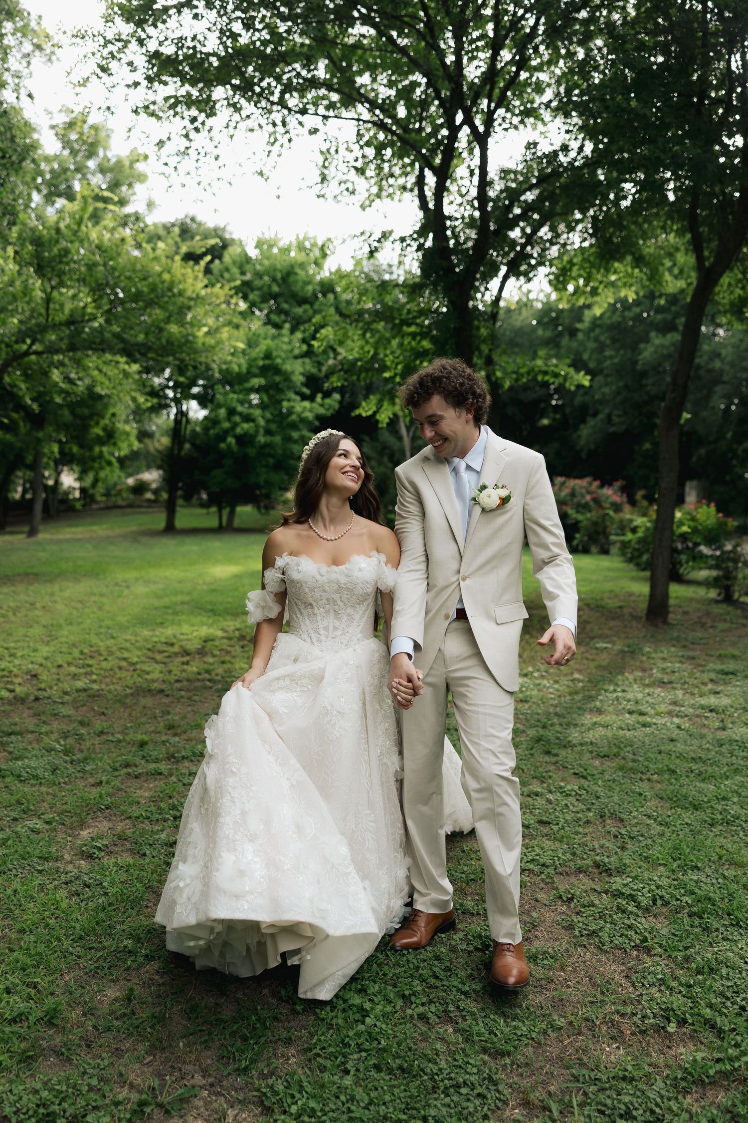A smiling bride in a white off-shoulder gown holds hands with a groom in a beige suit as they walk through a lush green park.
