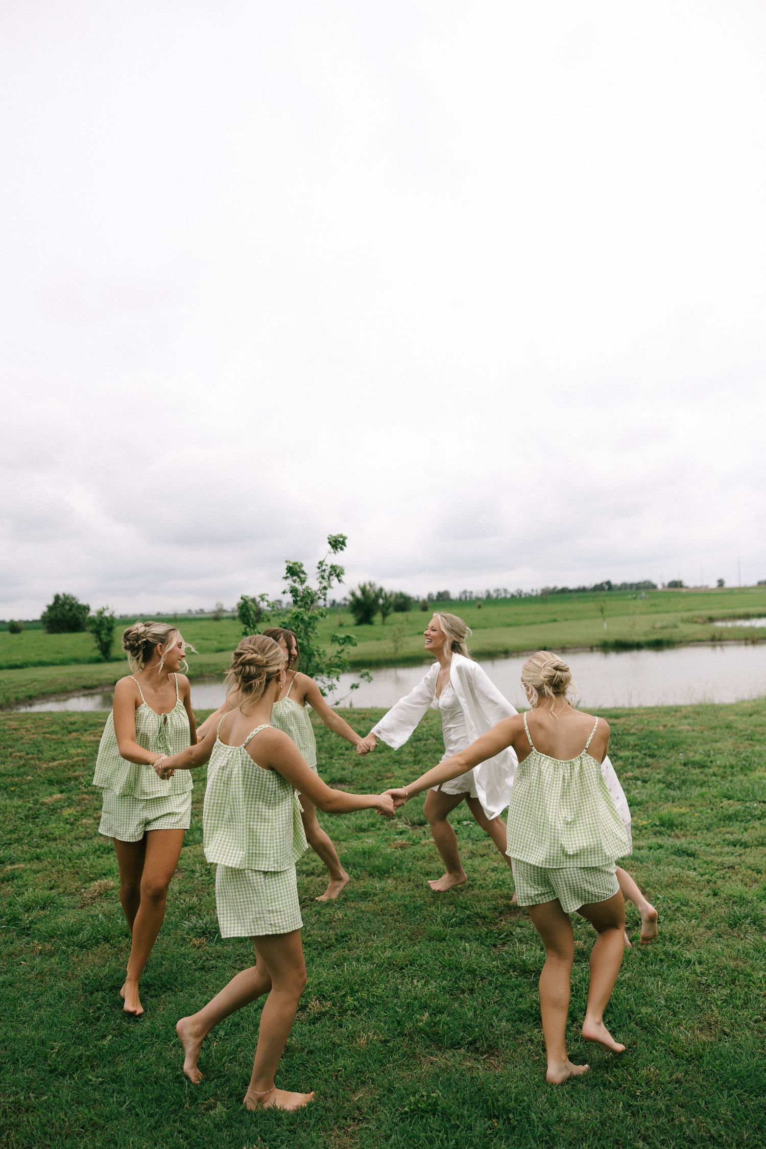 Bridesmaids in matching green pajamas dance in a circle barefoot on grass near a pond with a bride in white.