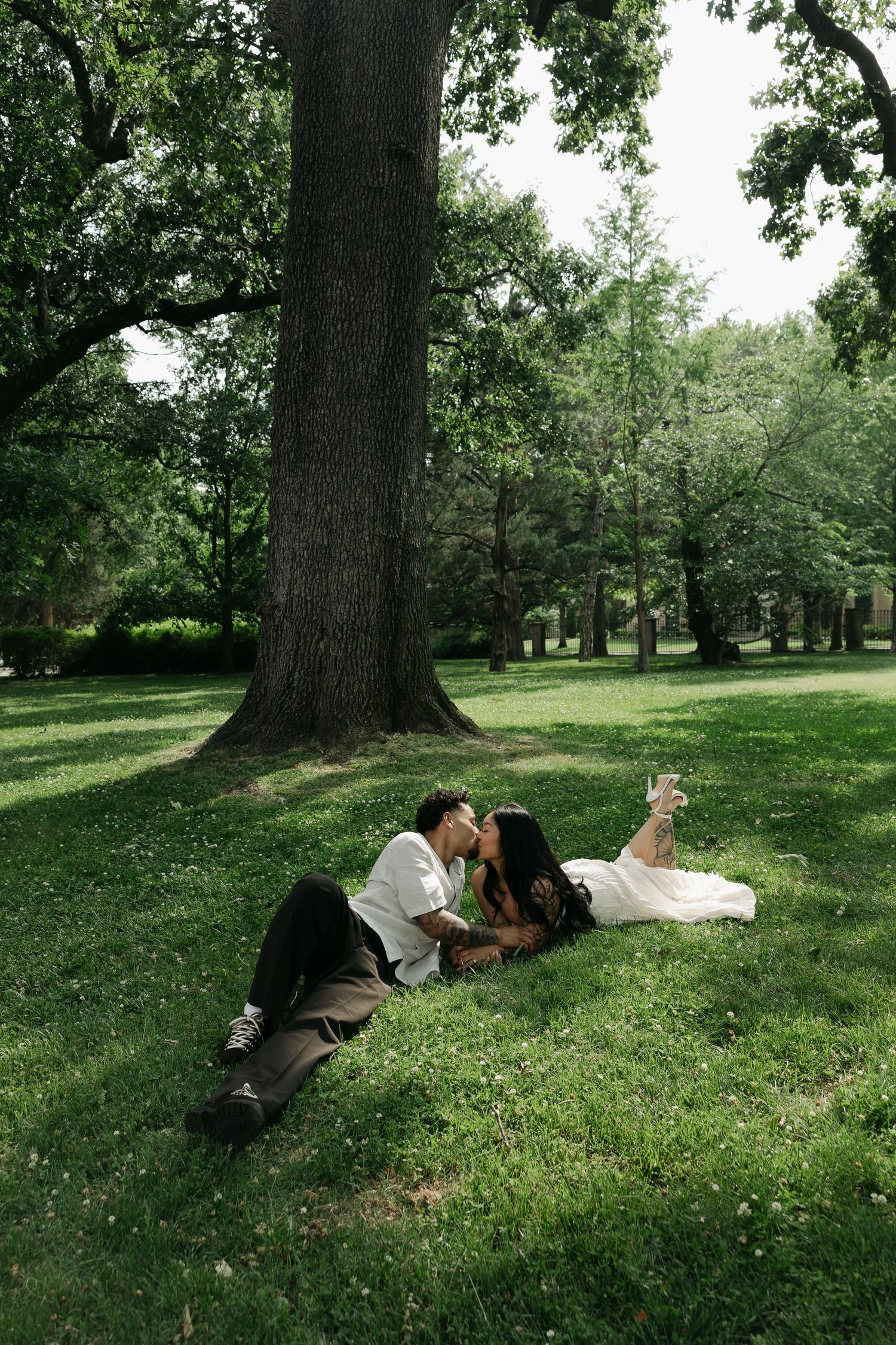 Bride and groom kissing while lying on lush green grass beneath a large tree in a park.