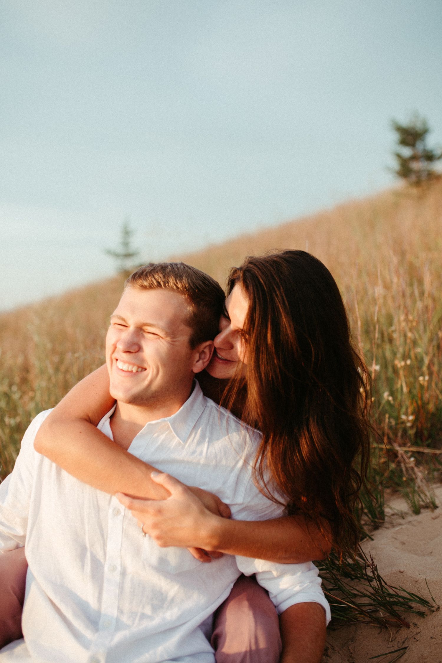 Woman kisses mans cheek as he laughs while sitting together on sandy dunes.