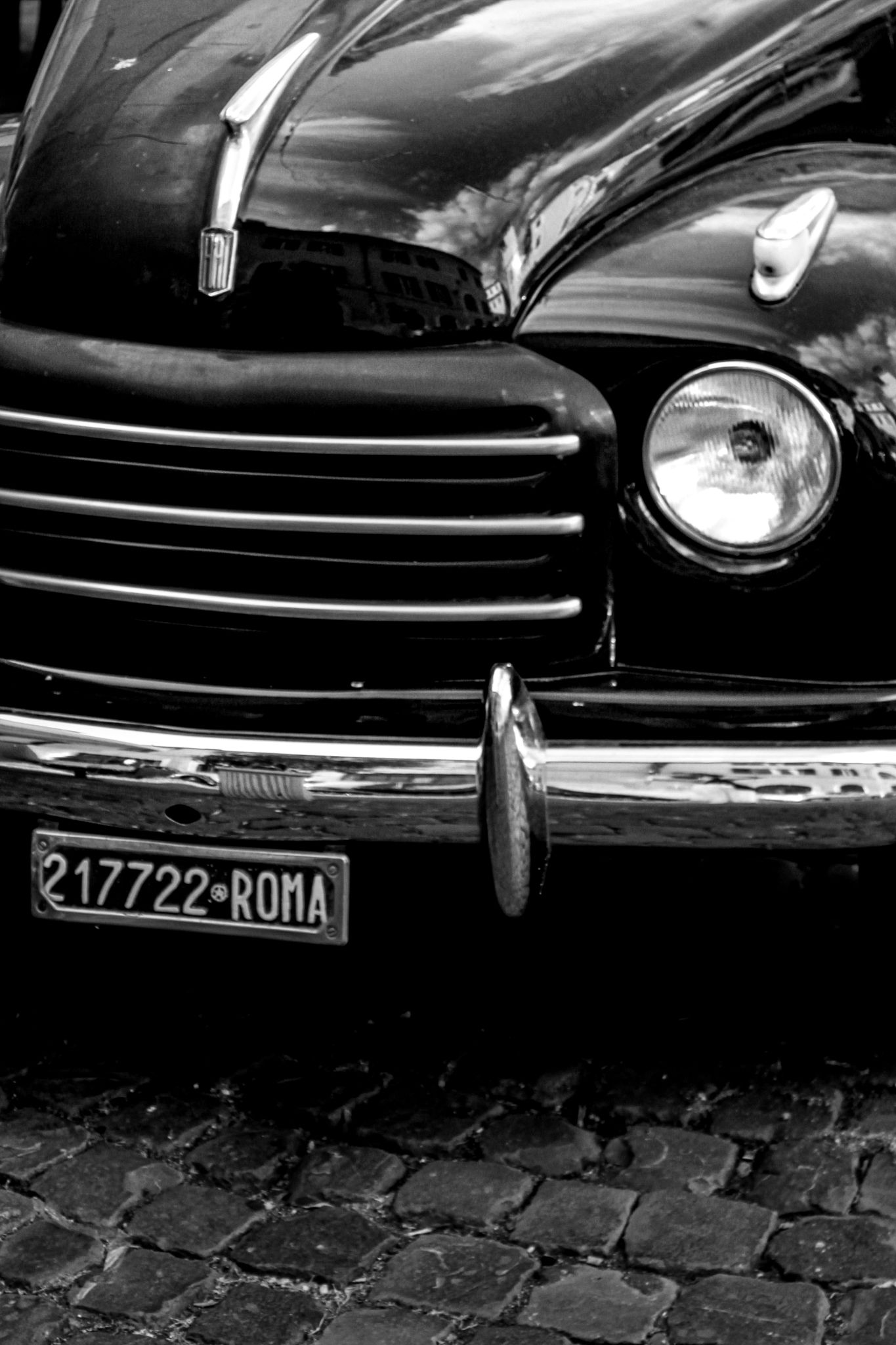 Black and white photograph of a vintage car with a Roma license plate on a cobblestone street in Rome