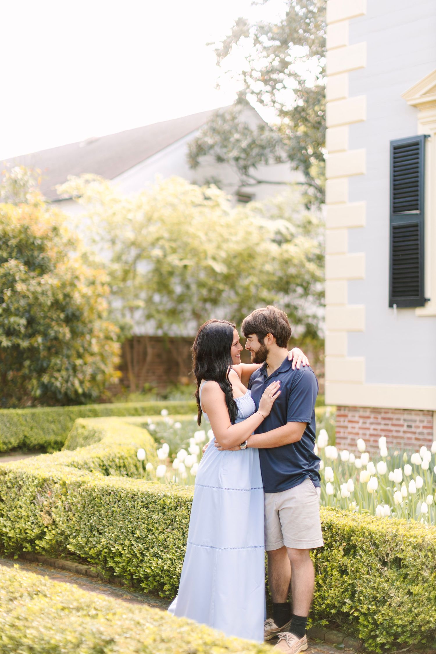 Couple embracing in a formal garden with white tulips and manicured hedges beside a historic building.