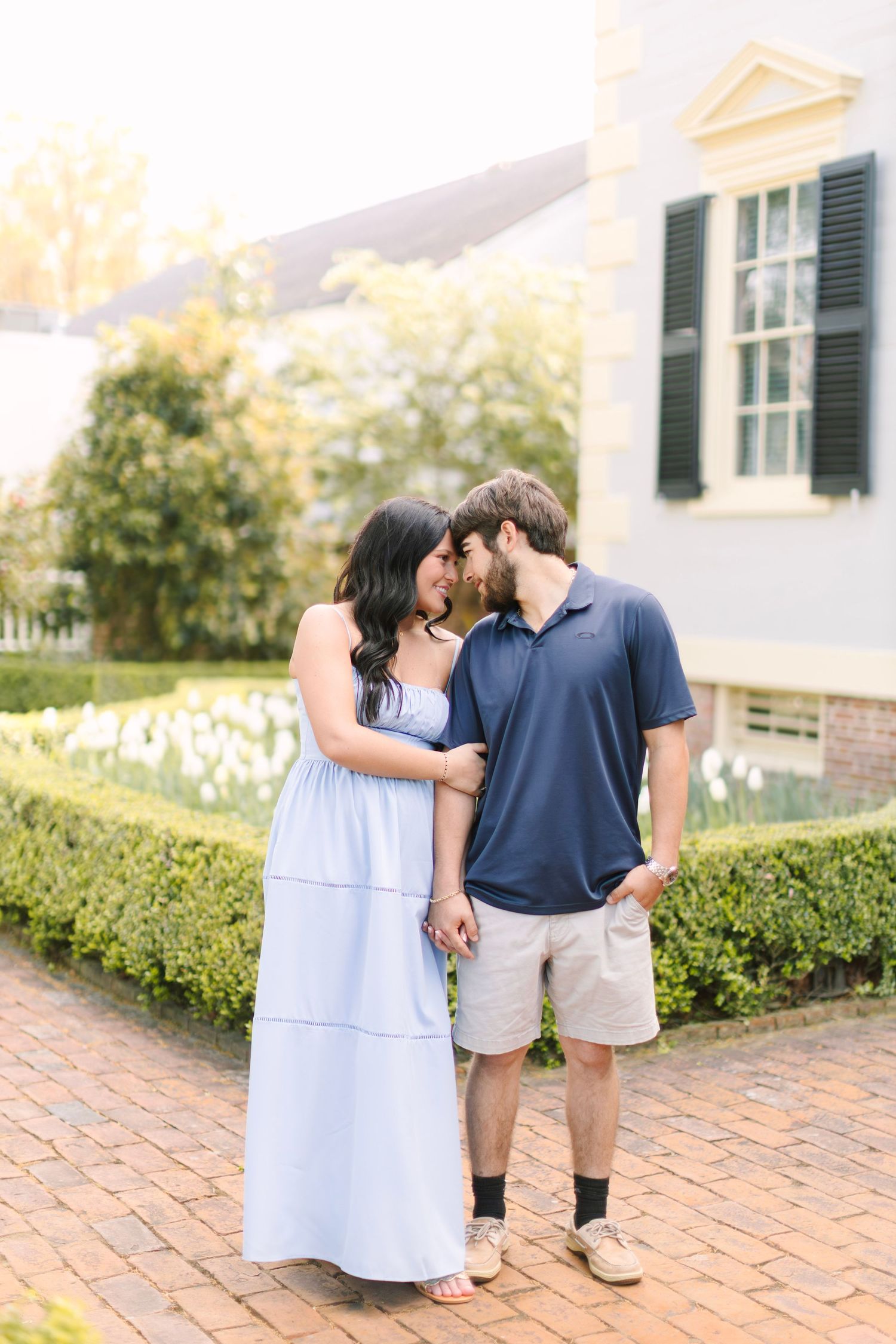 Couple sharing a tender moment on a brick path, woman in light blue maxi dress, man in navy polo and khaki shorts.