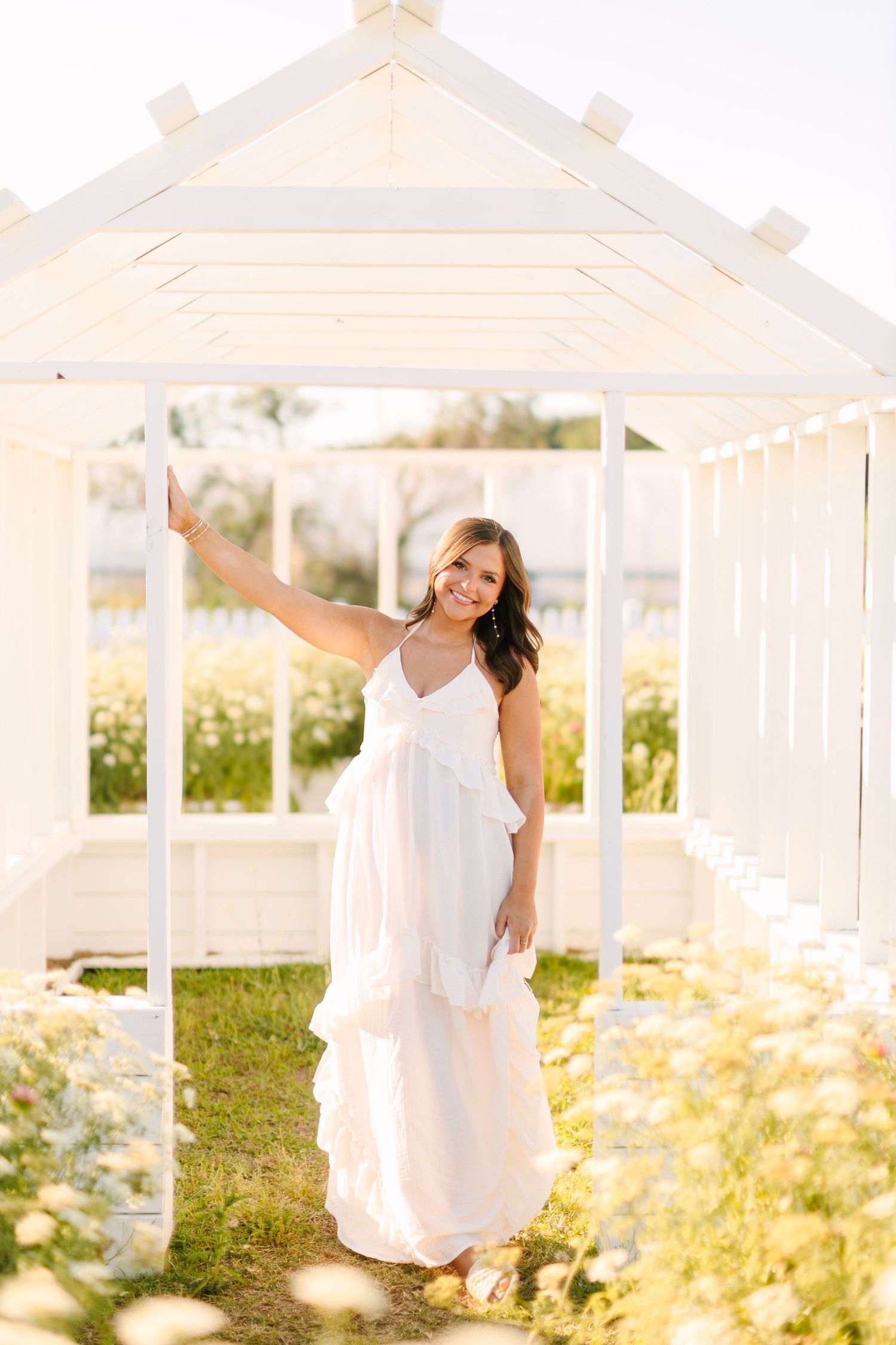 A smiling woman in a white ruffled maxi dress poses under a white pergola surrounded by wildflowers.
