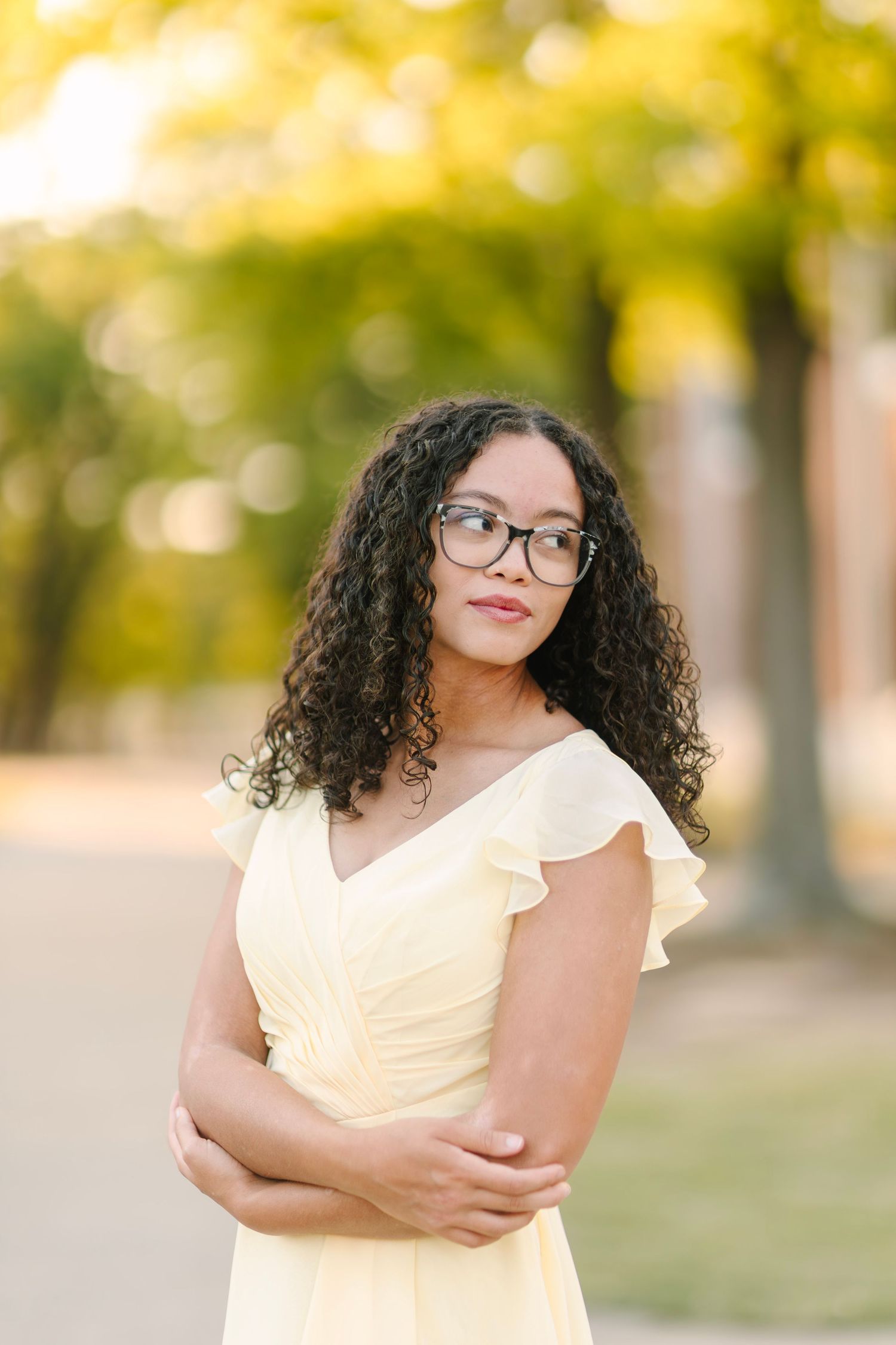 Young woman with curly hair and glasses in a yellow dress stands outdoors among autumn trees.
