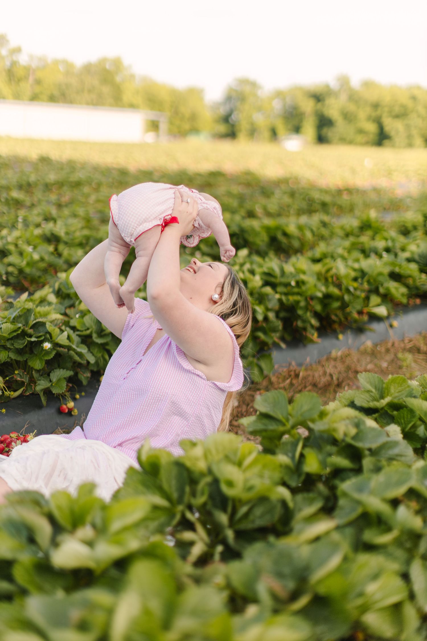 Mother lifting baby in strawberry field, both wearing pink dresses, sharing joyful moment among lush green plants.