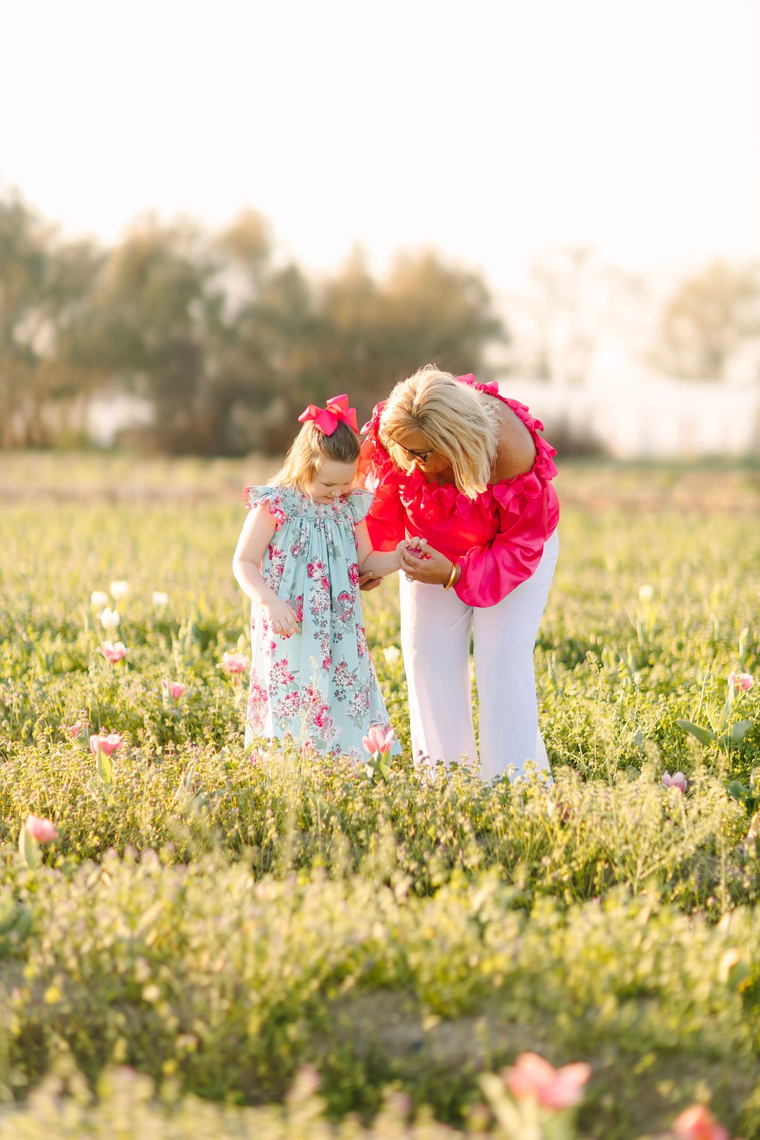 Woman in red top bends to show young girl in floral dress a flower in a sunny pink wildflower field.