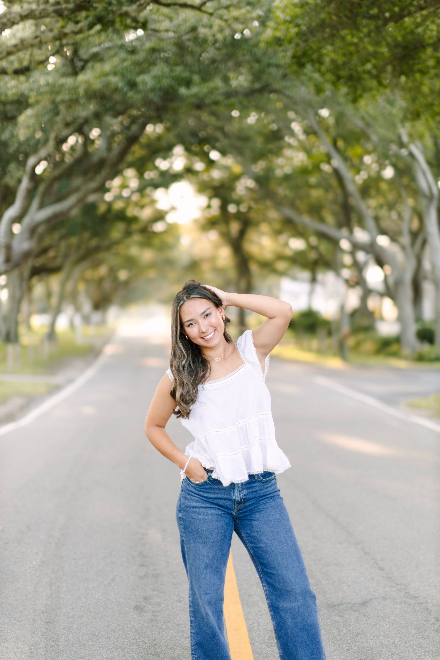 Young woman in white top and jeans poses on tree-lined road, smiling confidently in a bright outdoor setting.