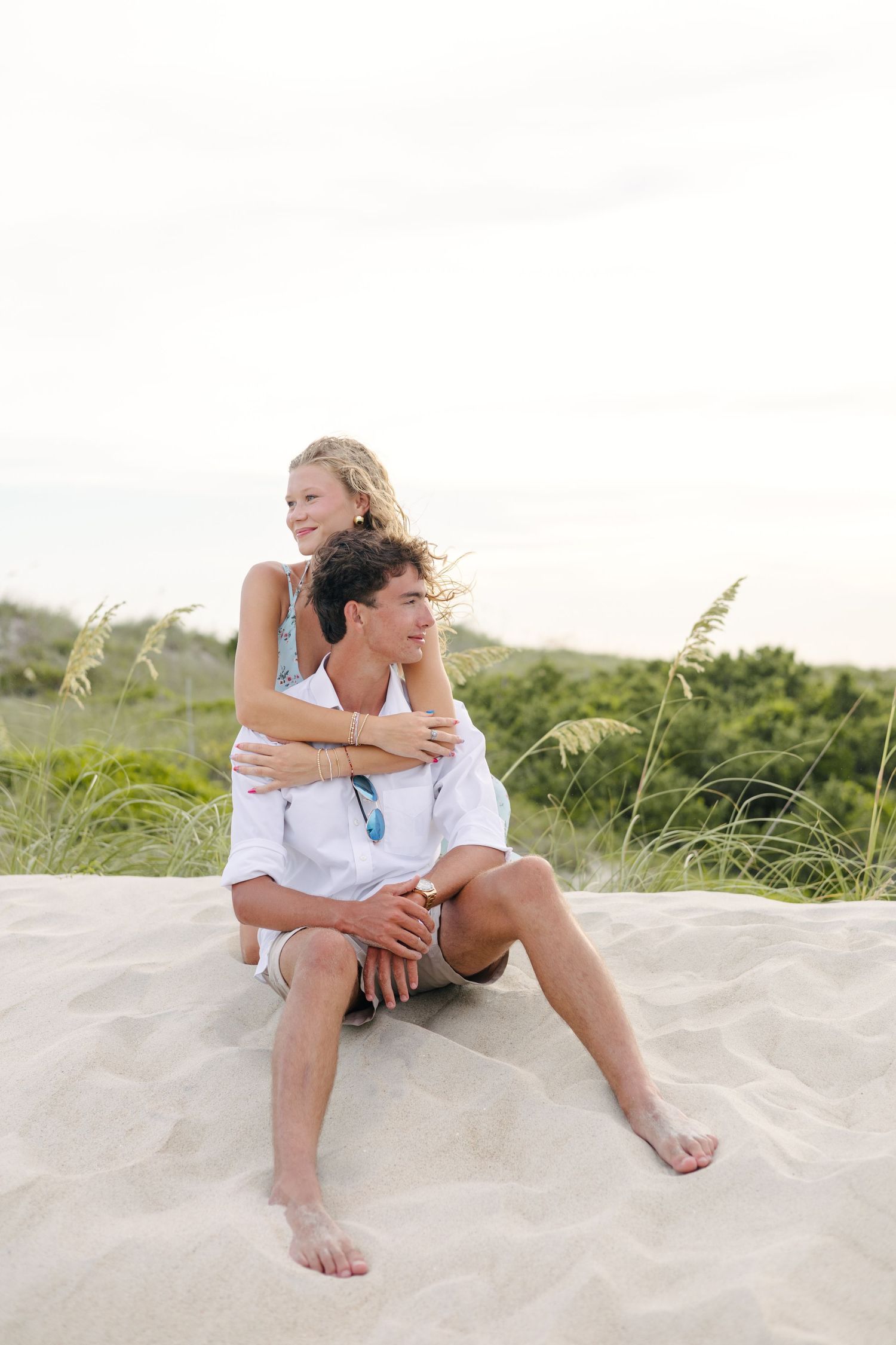 Couple shares intimate moment while sitting on white sand dunes at beach.