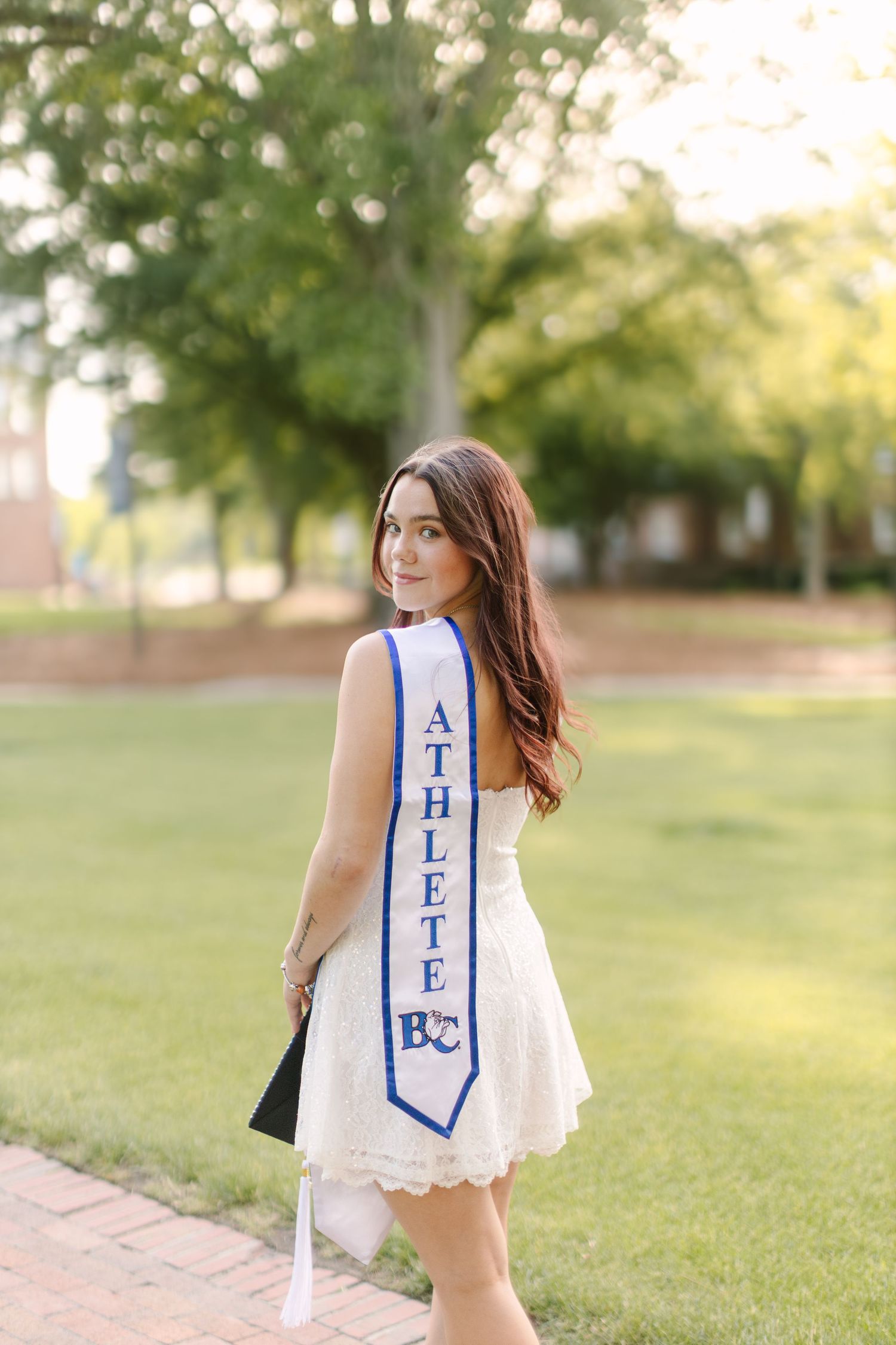 Young woman in white lace dress wearing an Athlete graduation sash poses outdoors on a sunny campus.