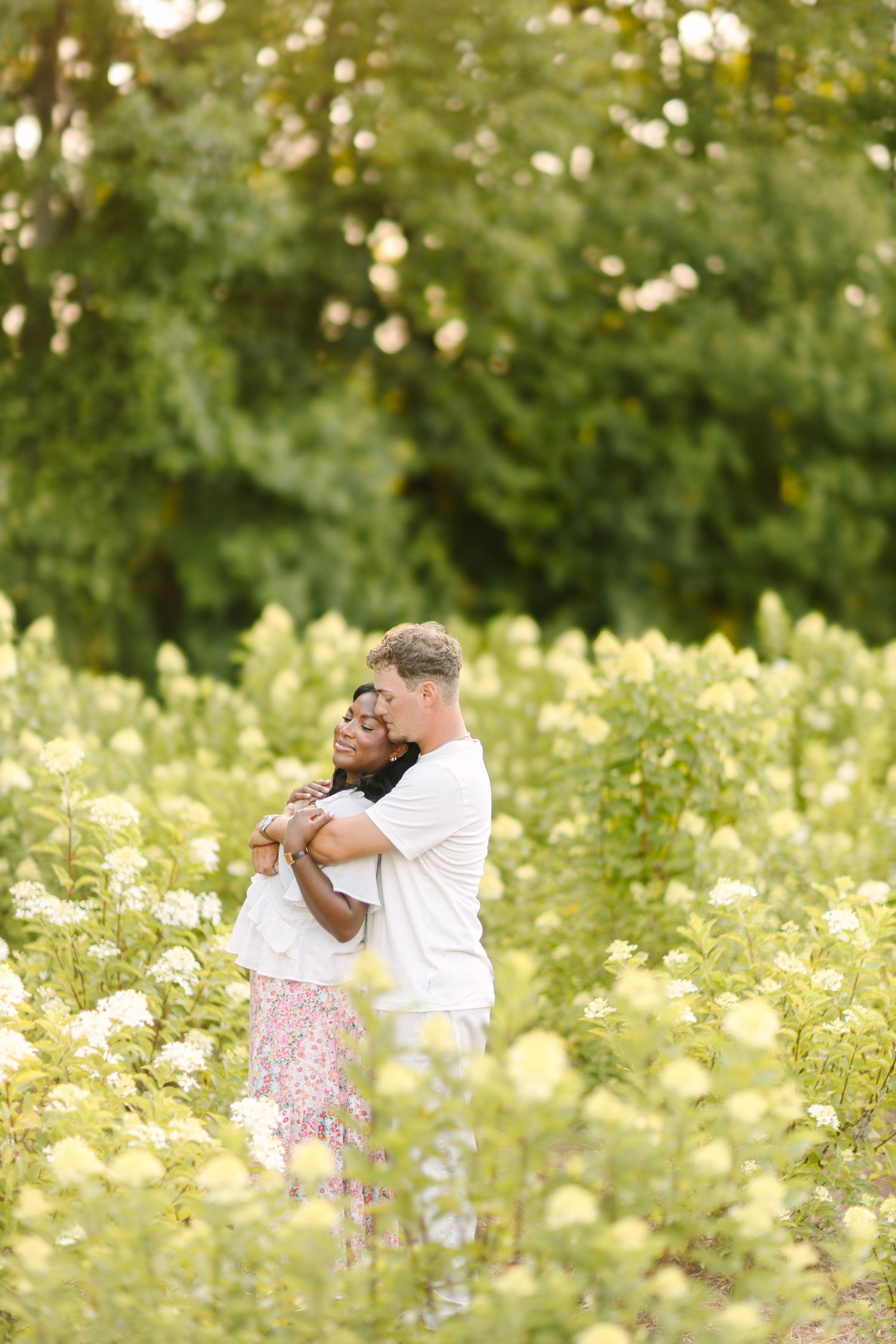 Couple embracing in a lush flower field with green trees, wearing white tops, sharing a romantic outdoor moment.