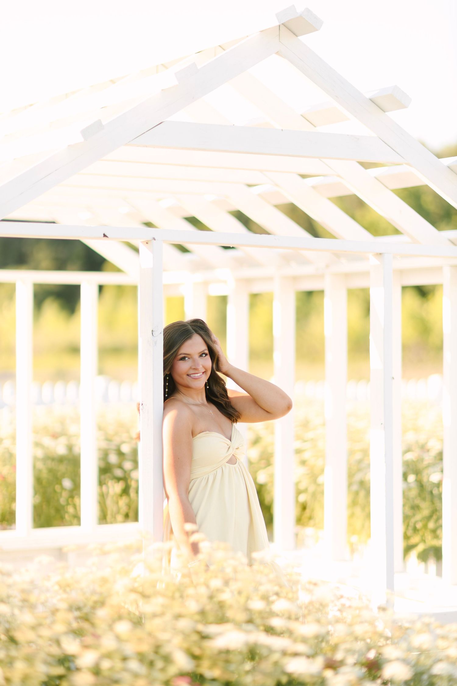 Young woman in a cream strapless dress smiling in a white pergola surrounded by wildflowers in golden sunlight.