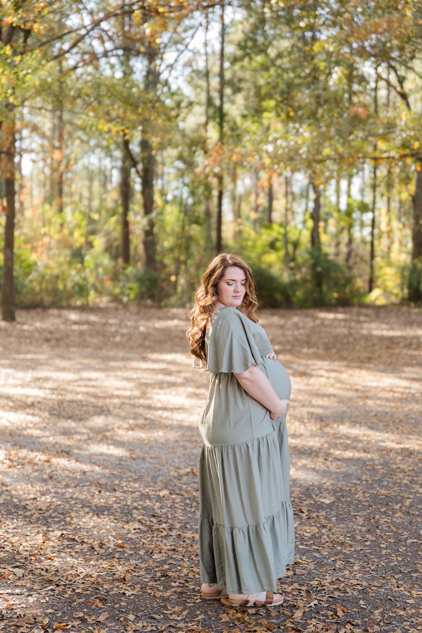 A person in a flowing sage green dress twirls in a sunlit forest clearing covered with fallen autumn leaves.