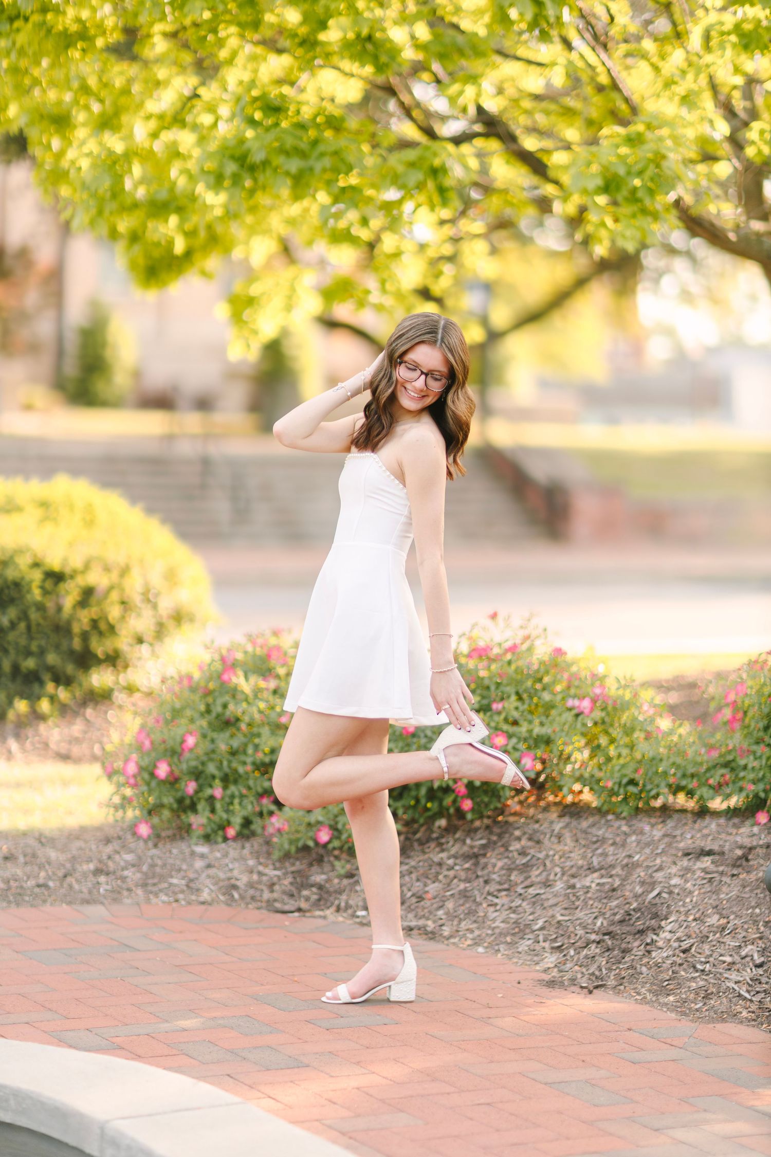 Young woman in white dress posing on brick path, lifting one leg, surrounded by colorful garden blooms and green trees.
