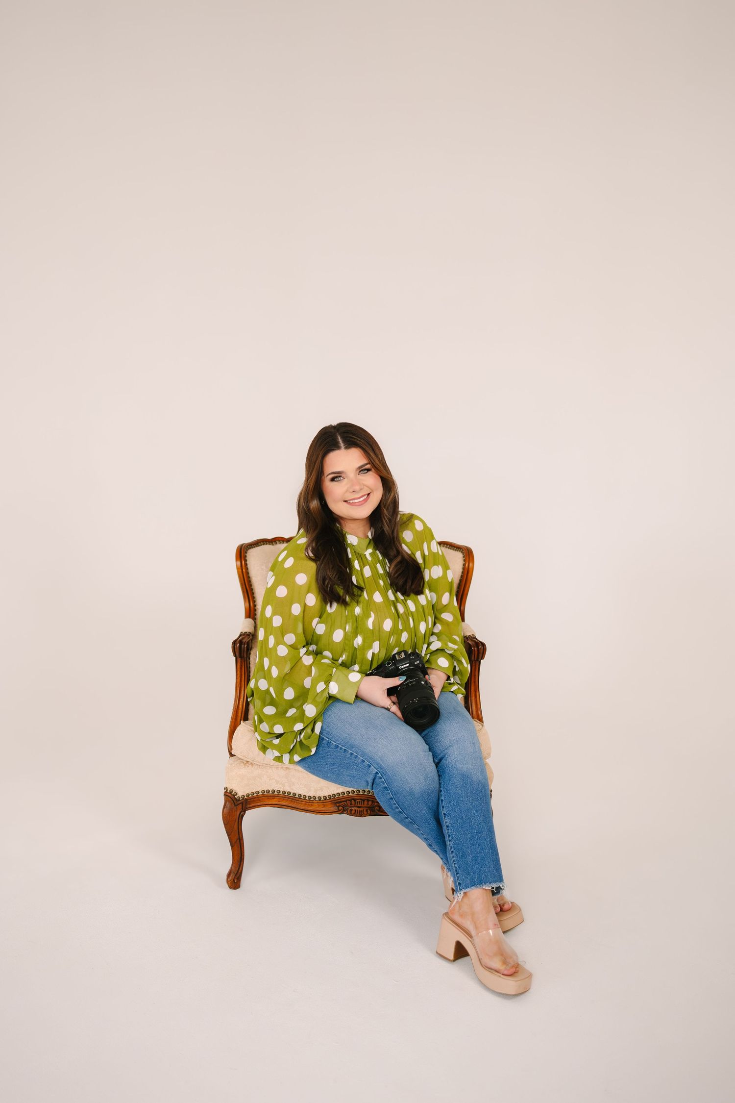 Woman in green polka dot blouse and jeans sits in ornate chair holding camera, smiling against neutral background.