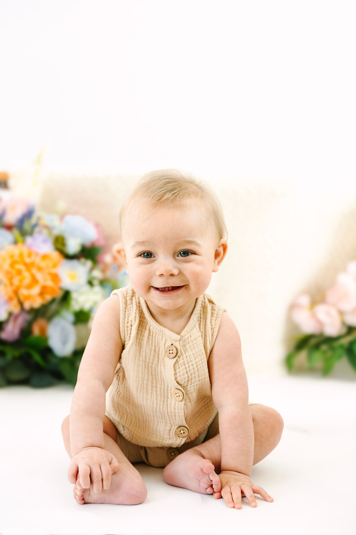 Smiling baby in beige romper sitting on white floor surrounded by colorful spring flowers.