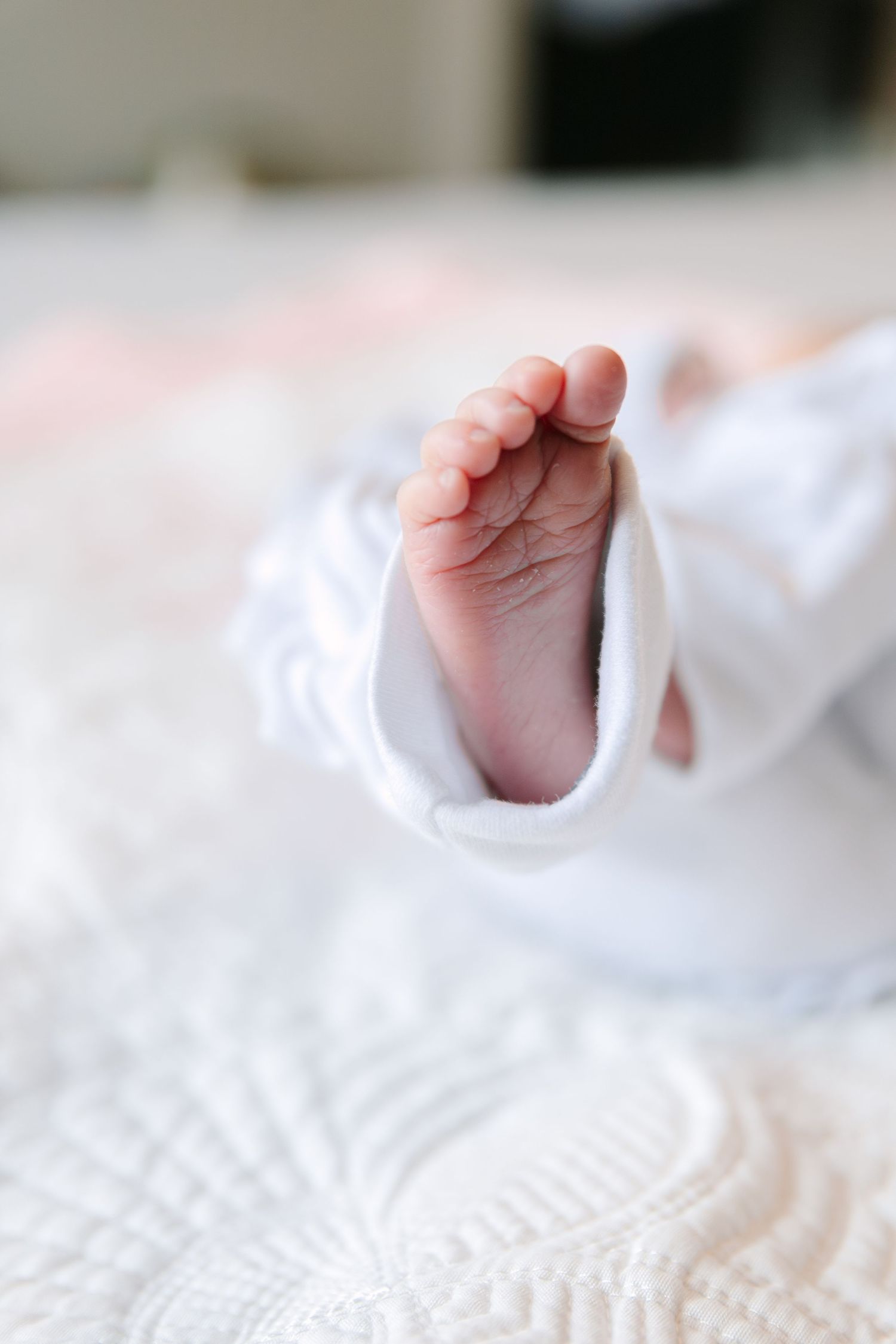 Close-up of a newborn baby's tiny foot peeking out from a white onesie, resting on a soft white blanket.