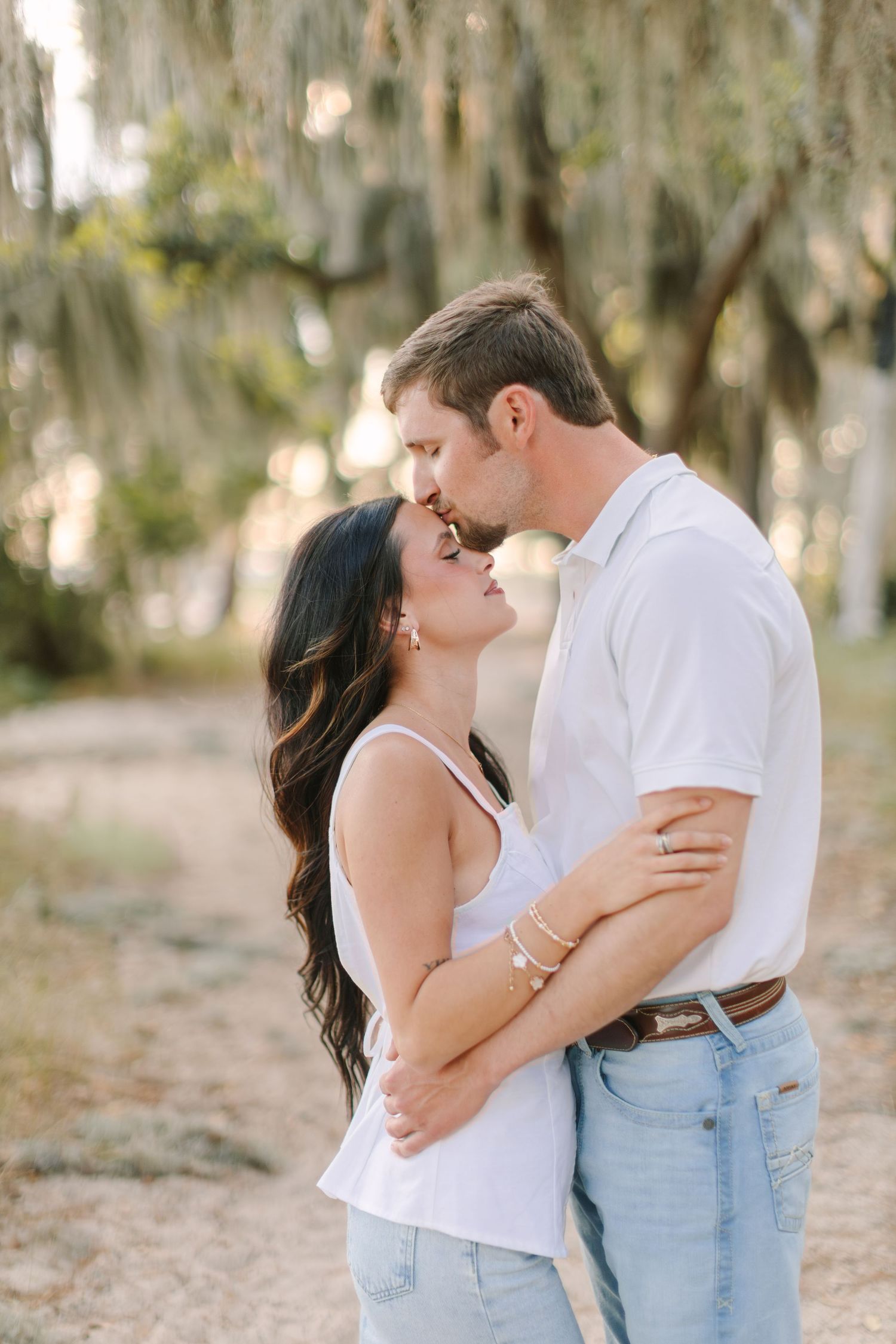 A couple shares a romantic kiss outdoors under Spanish moss-draped trees, dressed in white tops and light jeans.