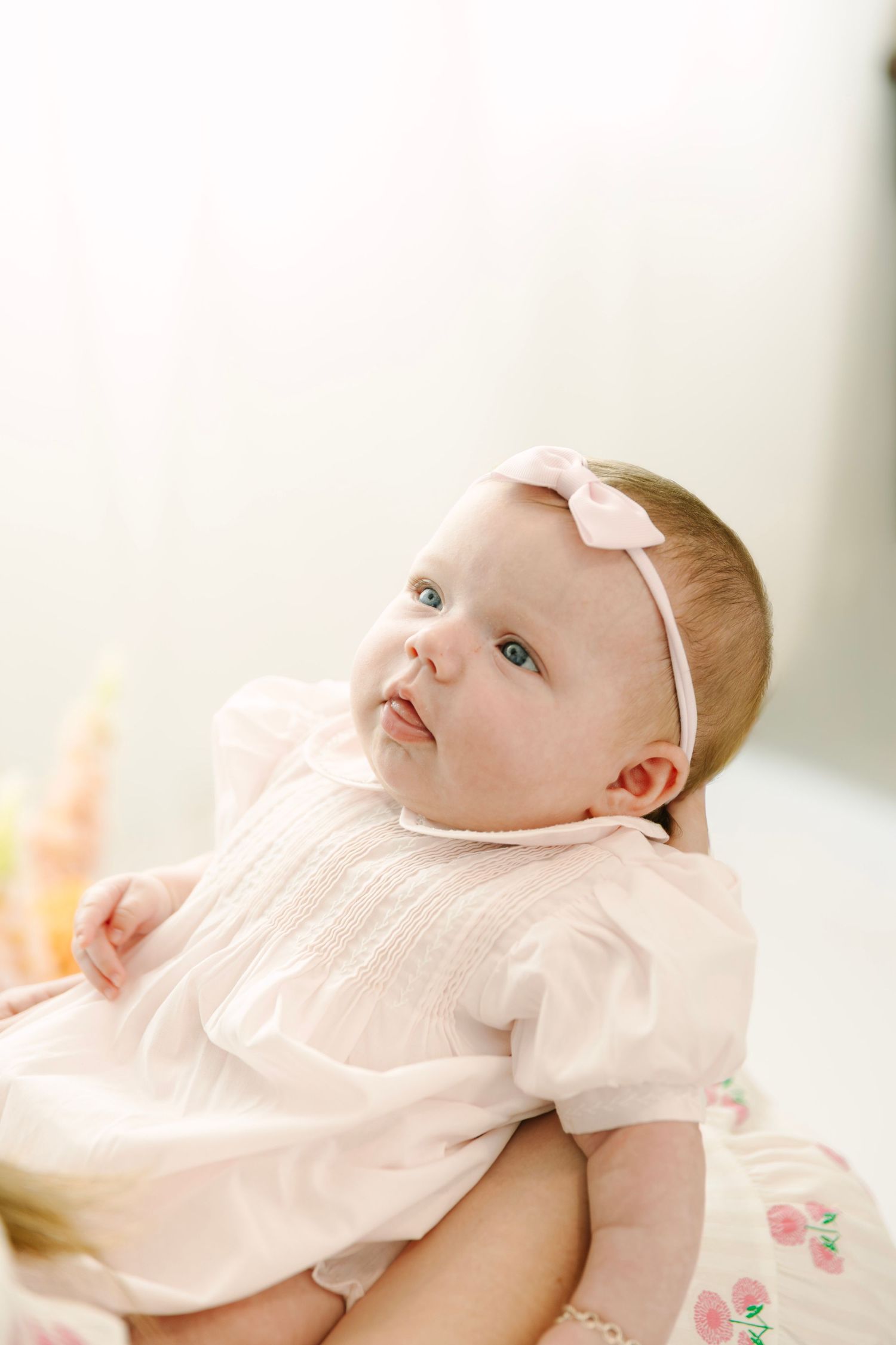 Adorable baby girl in pink dress and bow headband gazing upward with bright blue eyes.