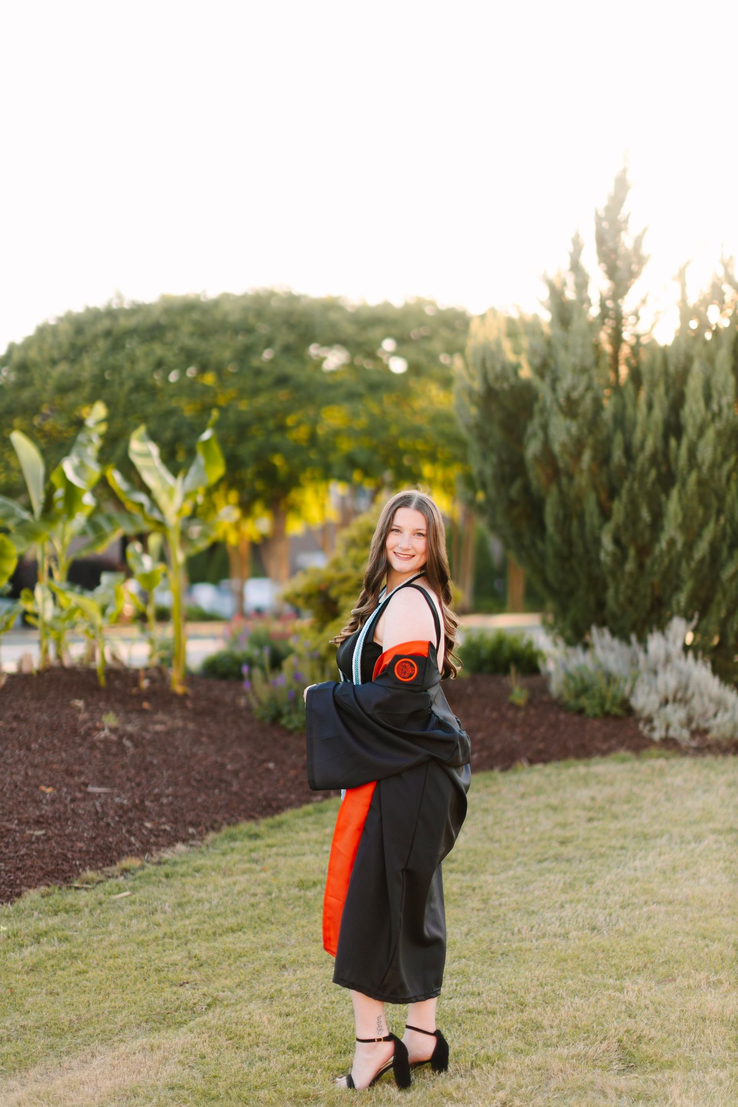 Smiling young woman in black graduation gown with orange accents posing outdoors on green lawn with trees in background.