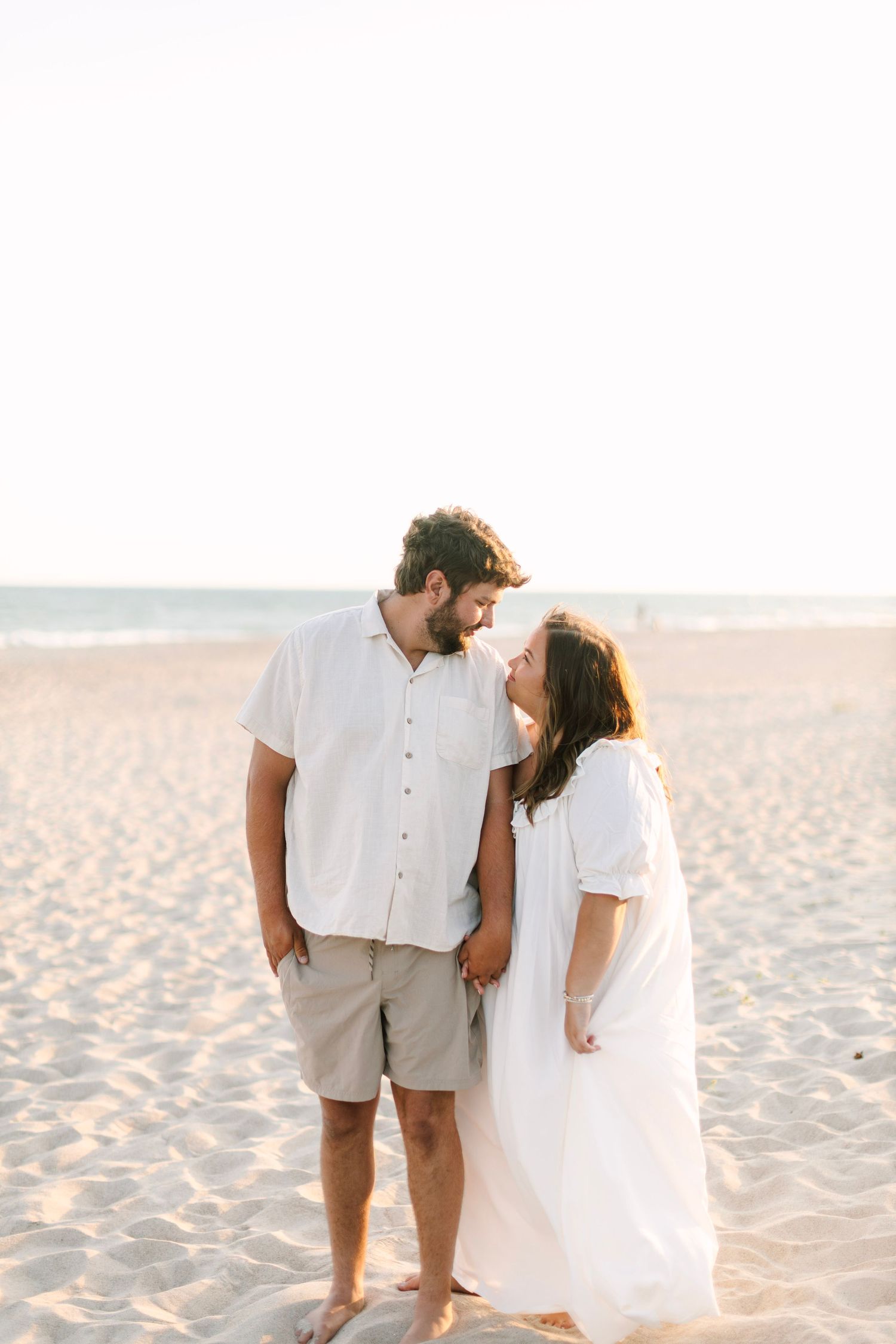 A couple in white outfits share a romantic moment on a sandy beach at golden hour.