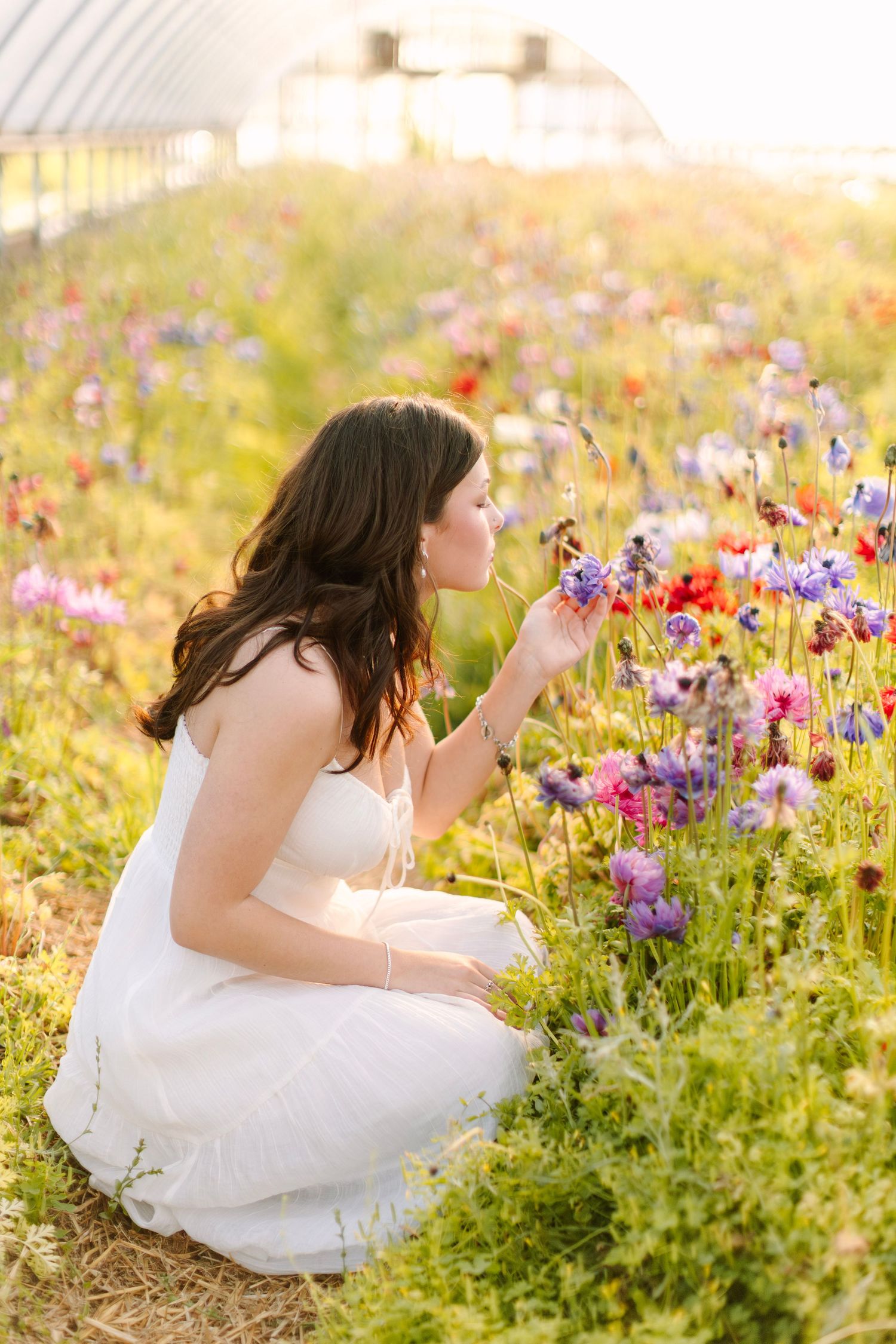 Woman in white dress crouching among colorful wildflowers in a sunlit greenhouse garden.