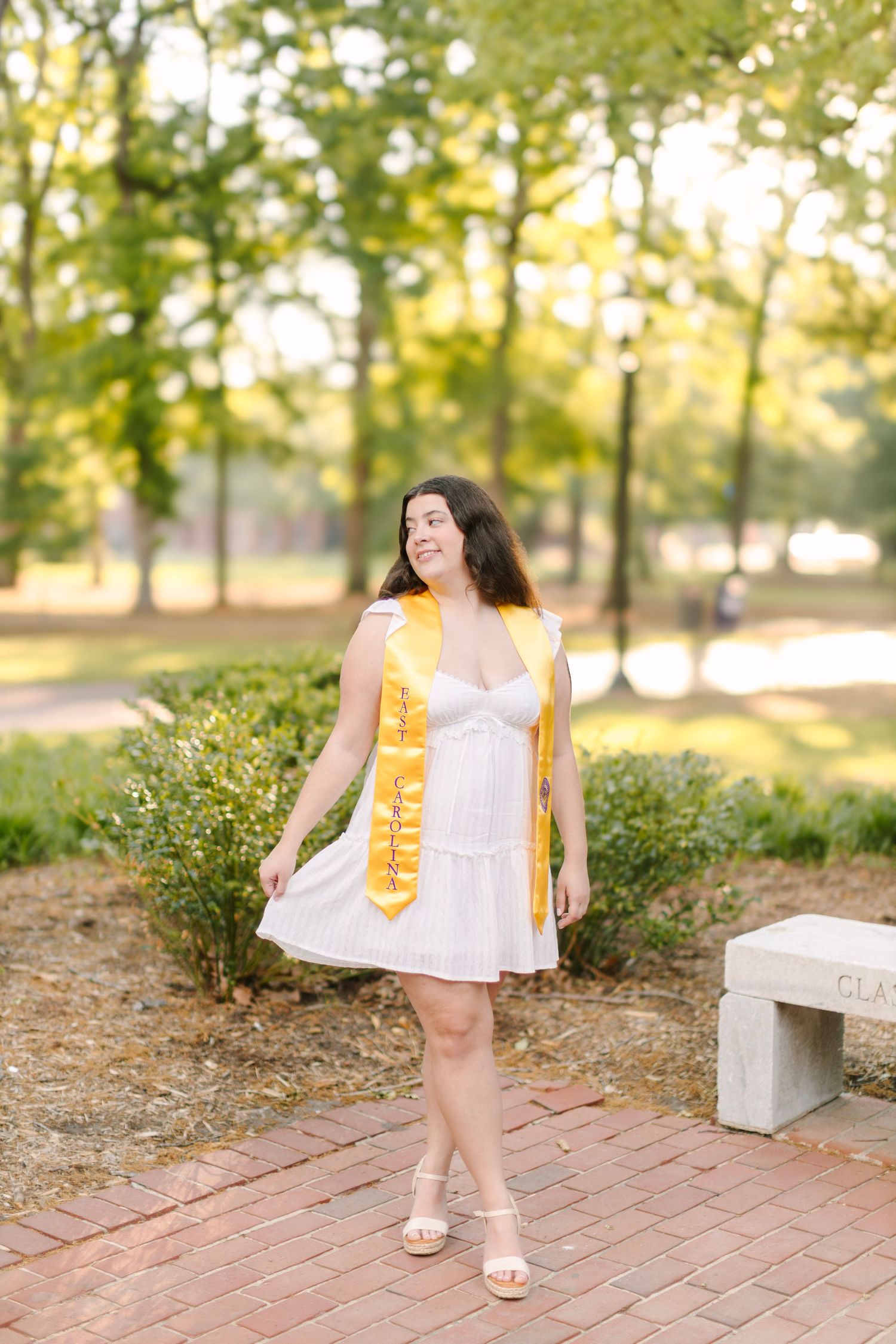 Young woman in white dress and gold graduation stole posing on brick path in a sunny park setting.