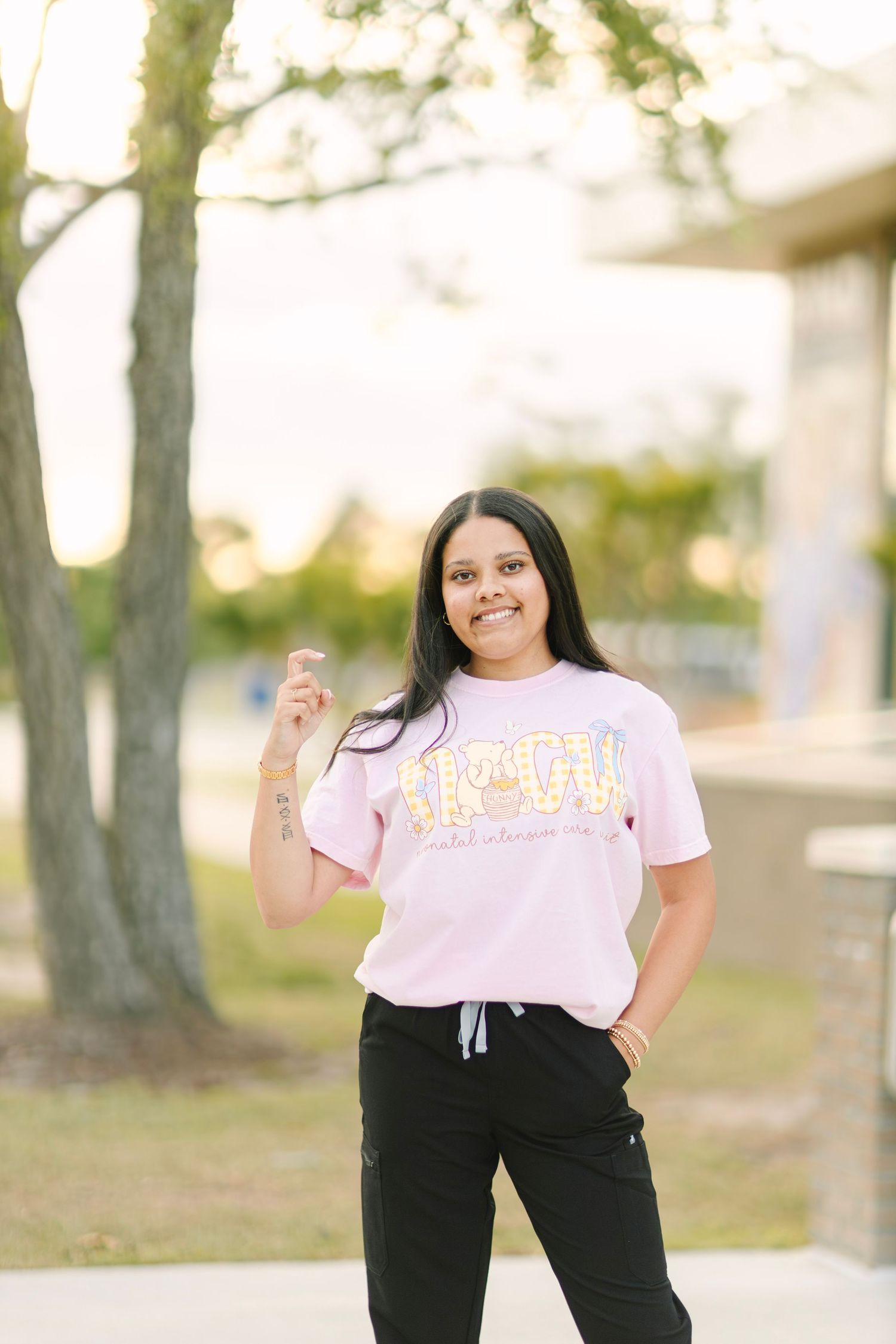 Young woman in pink HBCU graphic tee and black joggers posing outdoors near trees on a sunny day.