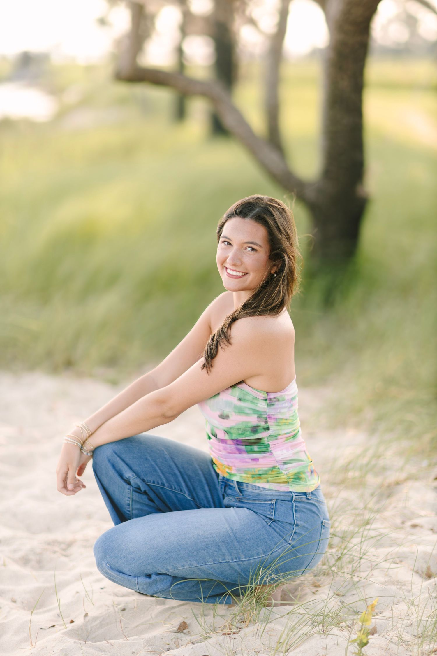 Smiling woman in colorful top and jeans sitting on sandy beach dunes with trees and green grass background.