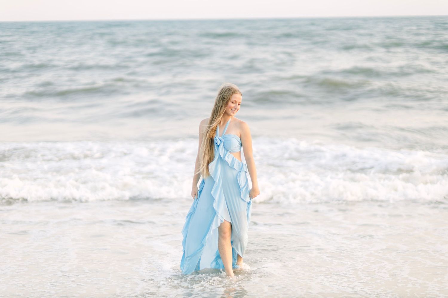 A woman in a flowing light blue dress walks gracefully along a sandy beach with ocean waves behind her.