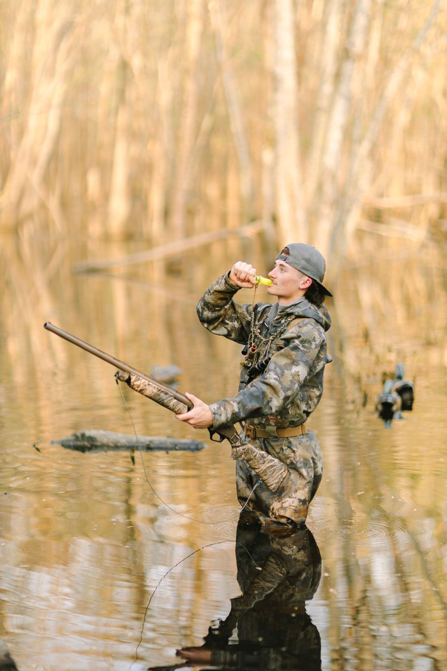 Hunter in camouflage wading through flooded forest, holding shotgun and blowing duck call during waterfowl hunting season.