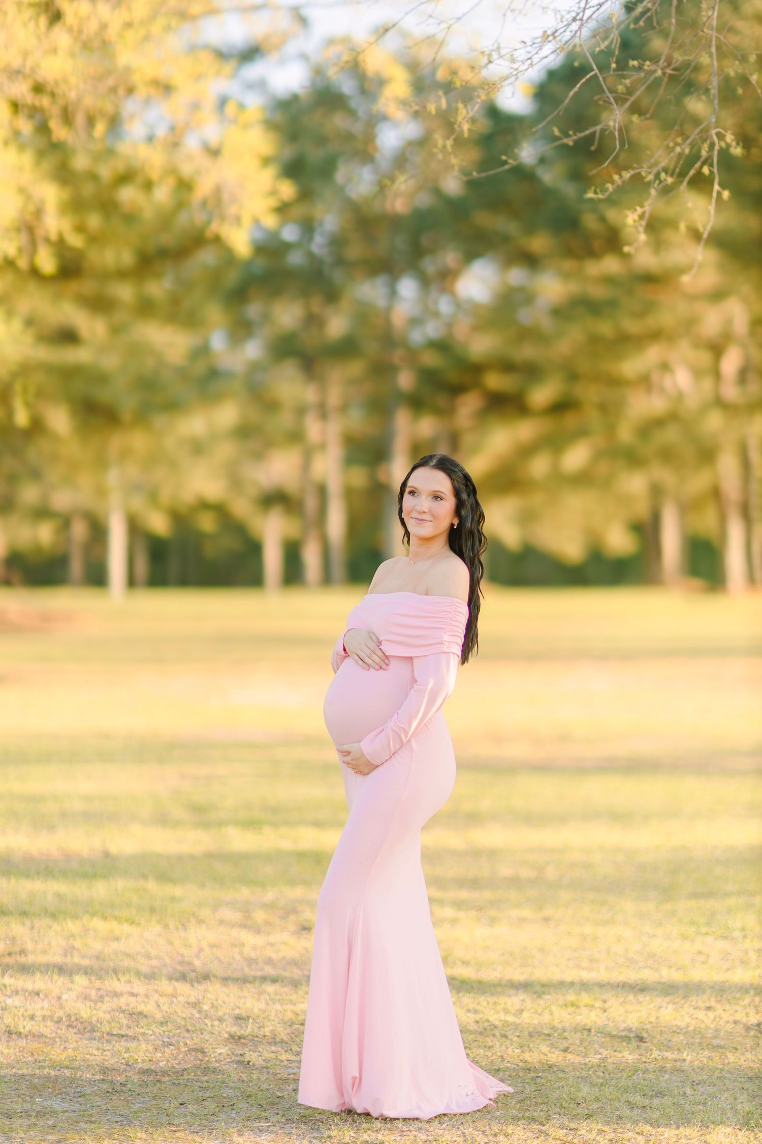 Pregnant woman in pink off-shoulder gown cradling baby bump outdoors in golden hour light.