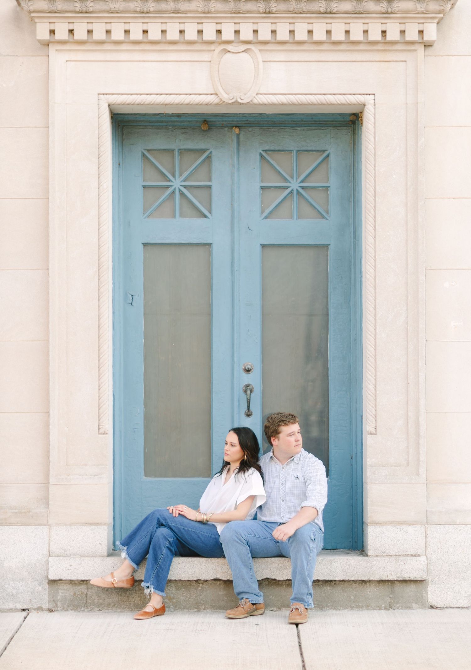 Couple sitting on steps in front of a blue double door at a classic stone building during an engagement session.