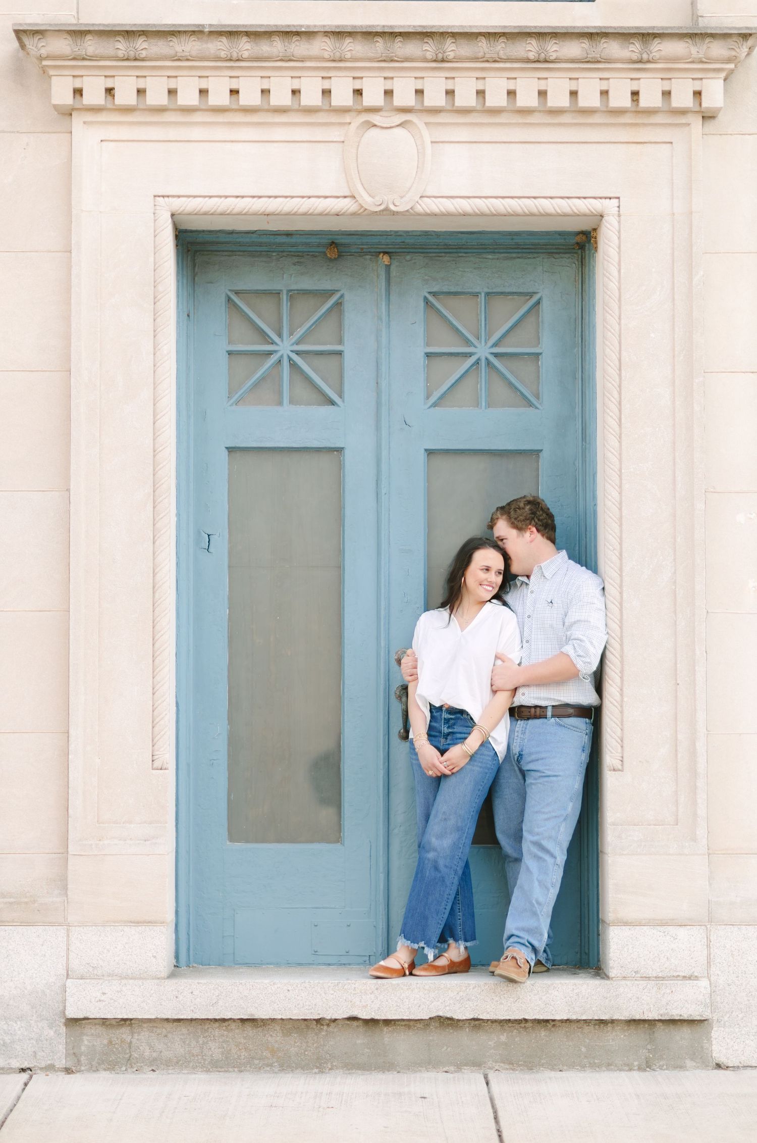 Couple leans against a blue door in a stone archway, woman in white top and jeans, man in light blue shirt.