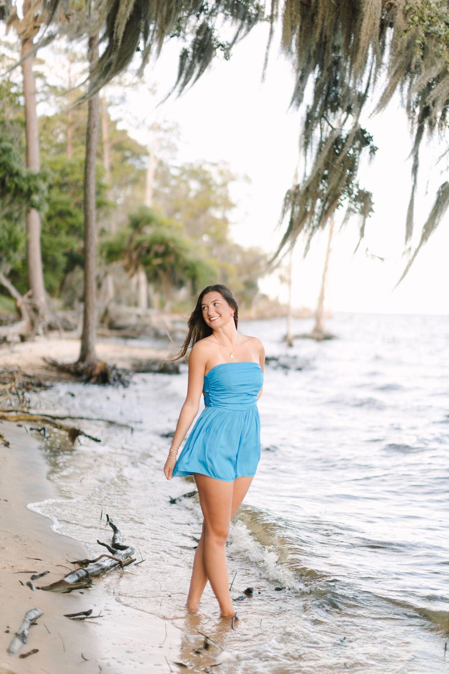 Young woman in blue strapless romper walking along a sandy beach shoreline beneath moss-draped trees.