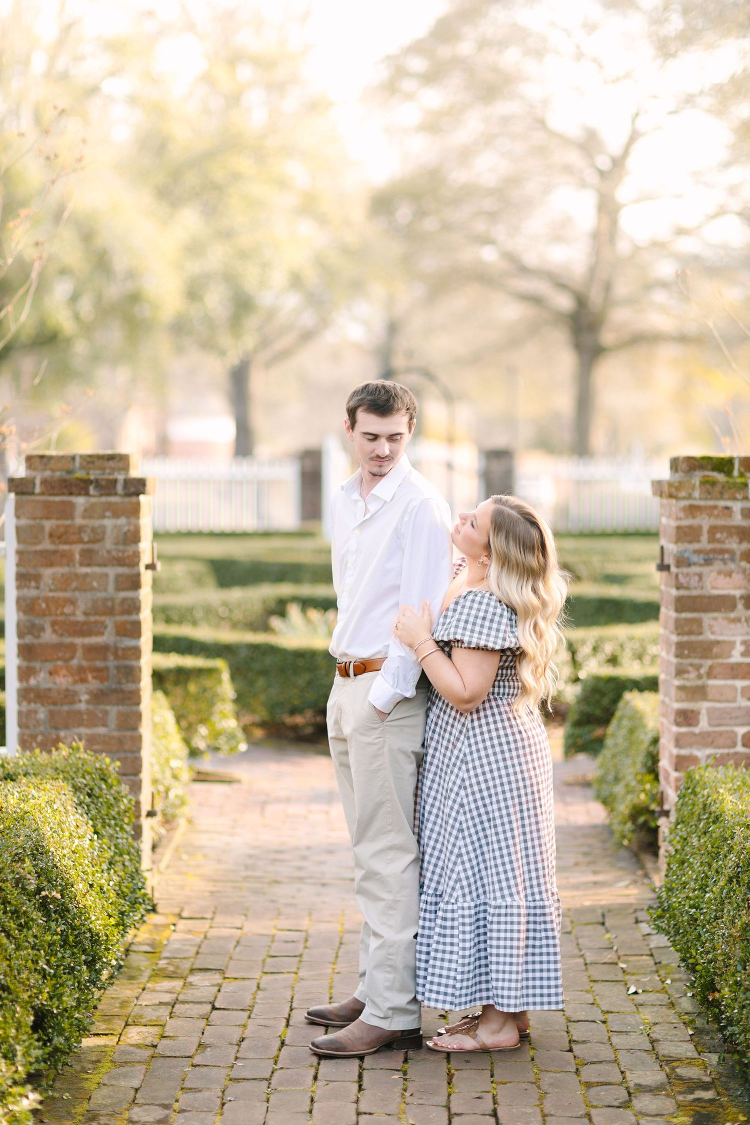 Couple embracing on brick path in formal garden, woman in gingham dress, man in white shirt, golden hour light.