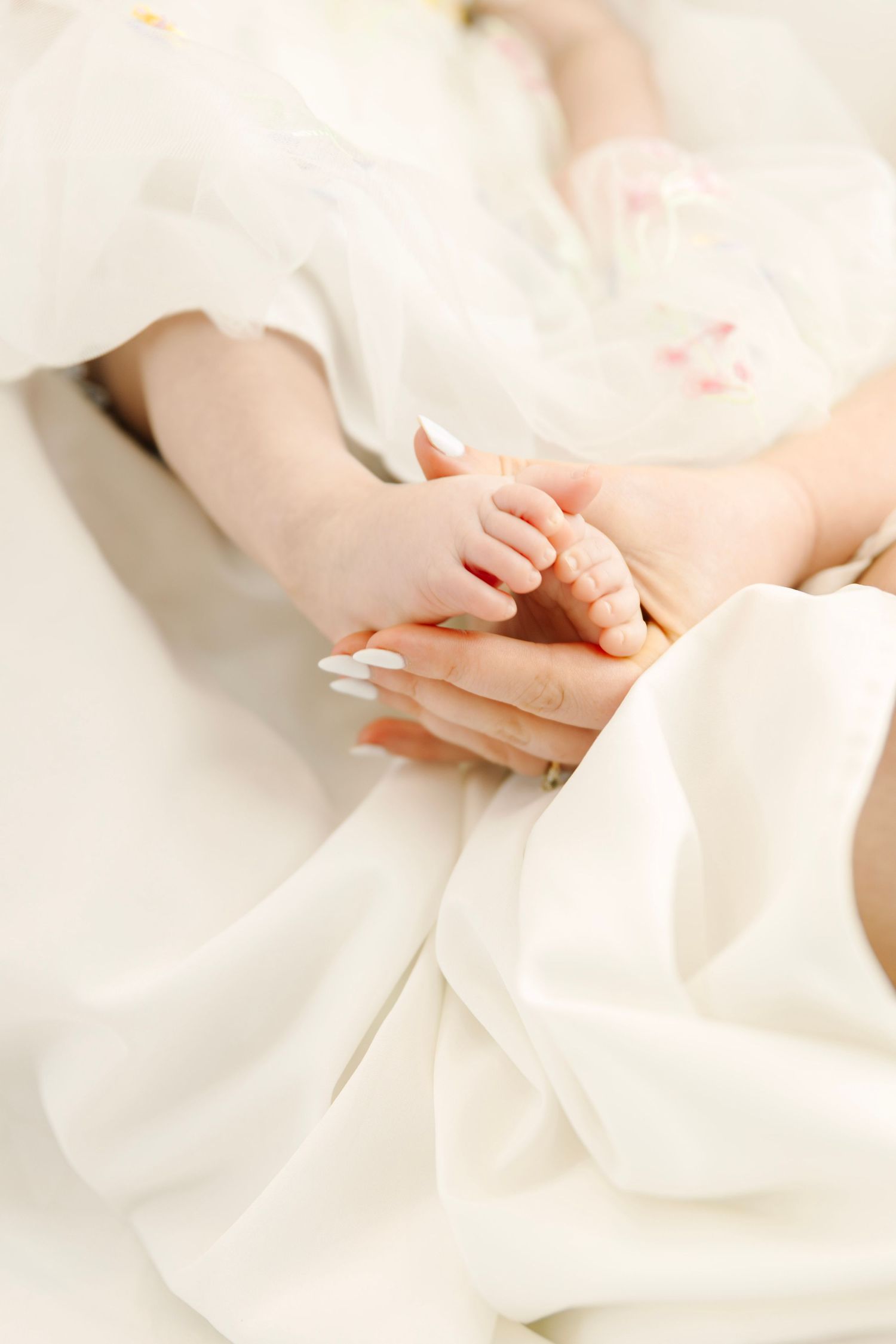 A mother's hand with white manicure gently holds her newborn baby's tiny hand on soft white fabric.