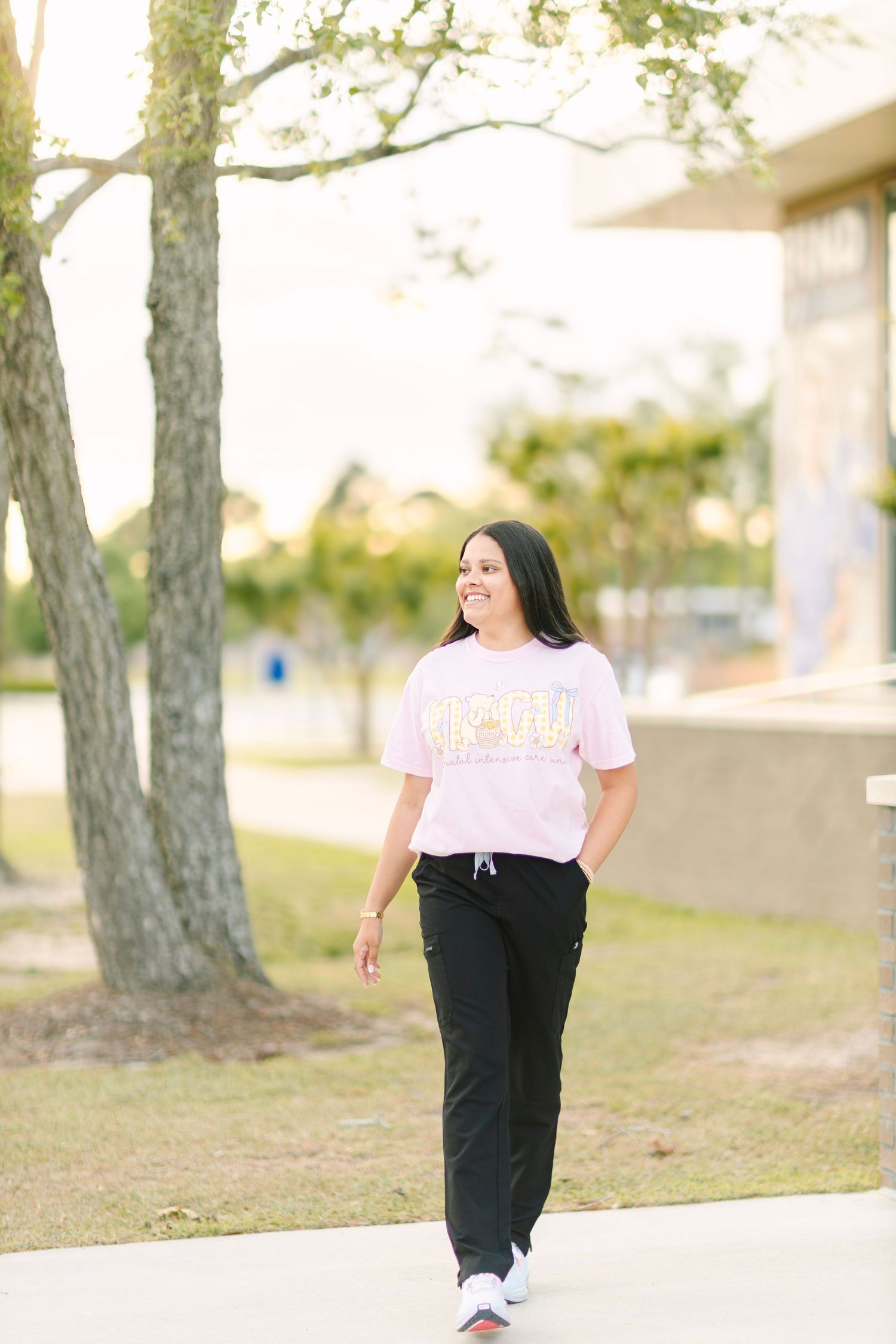 Woman in pink graphic tee and black scrub pants walking outdoors on a sunny day, smiling confidently.