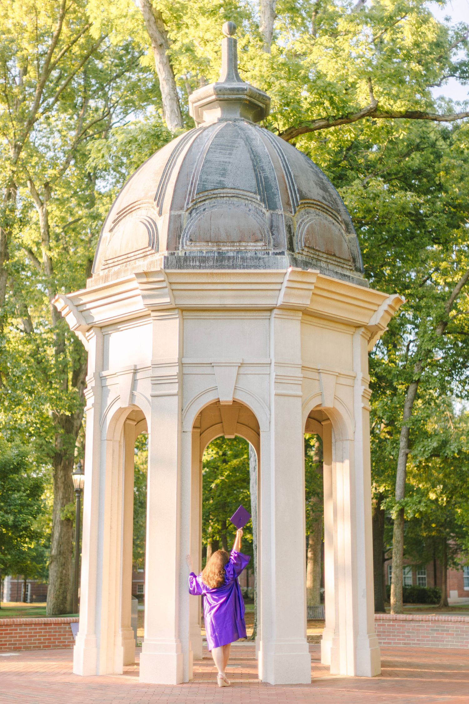 Graduate in purple gown celebrates under elegant domed gazebo surrounded by lush green trees on a sunny day.
