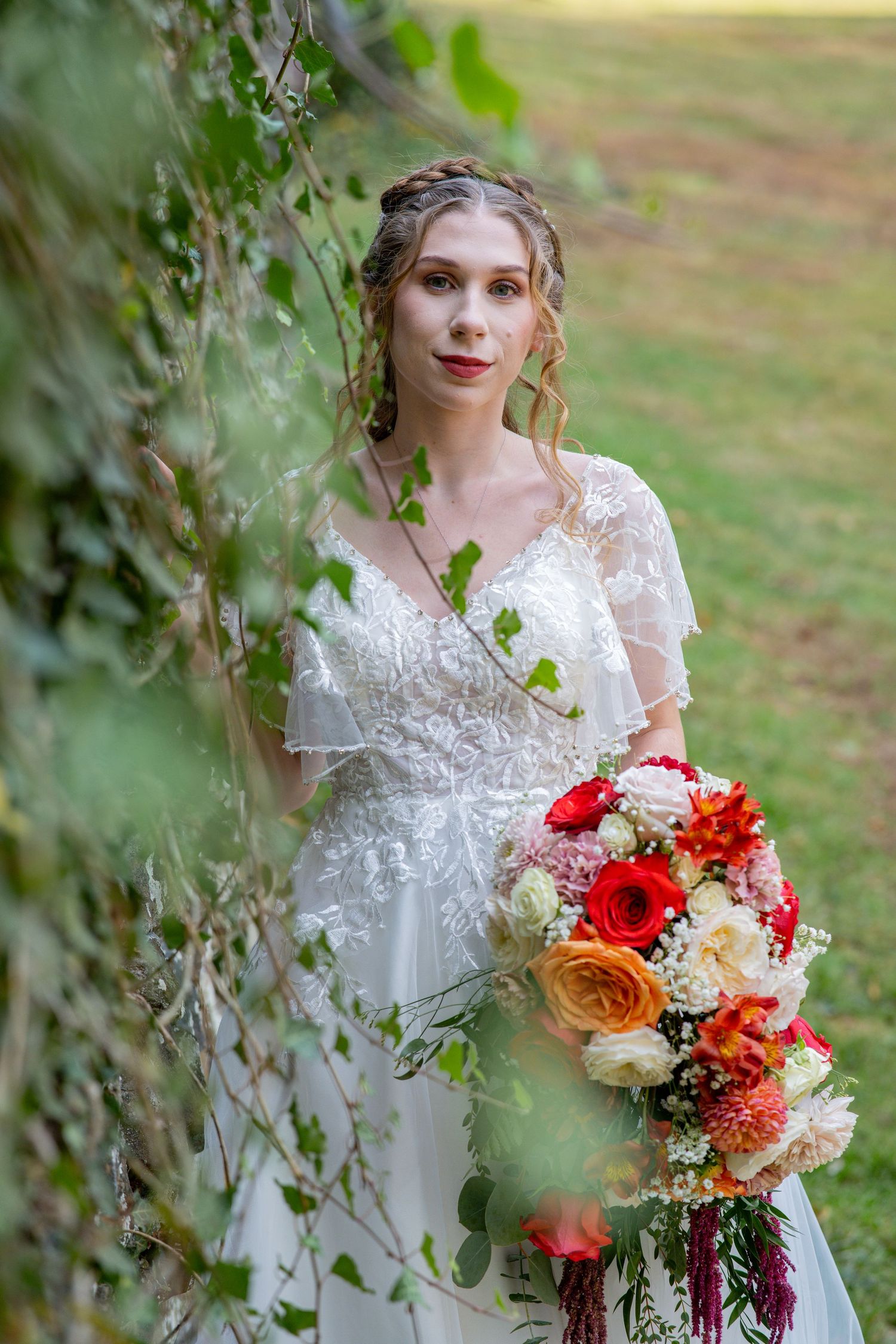 A bride in a delicate lace wedding dress holds a vibrant bouquet of red, orange and pink flowers against a garden backdrop.
