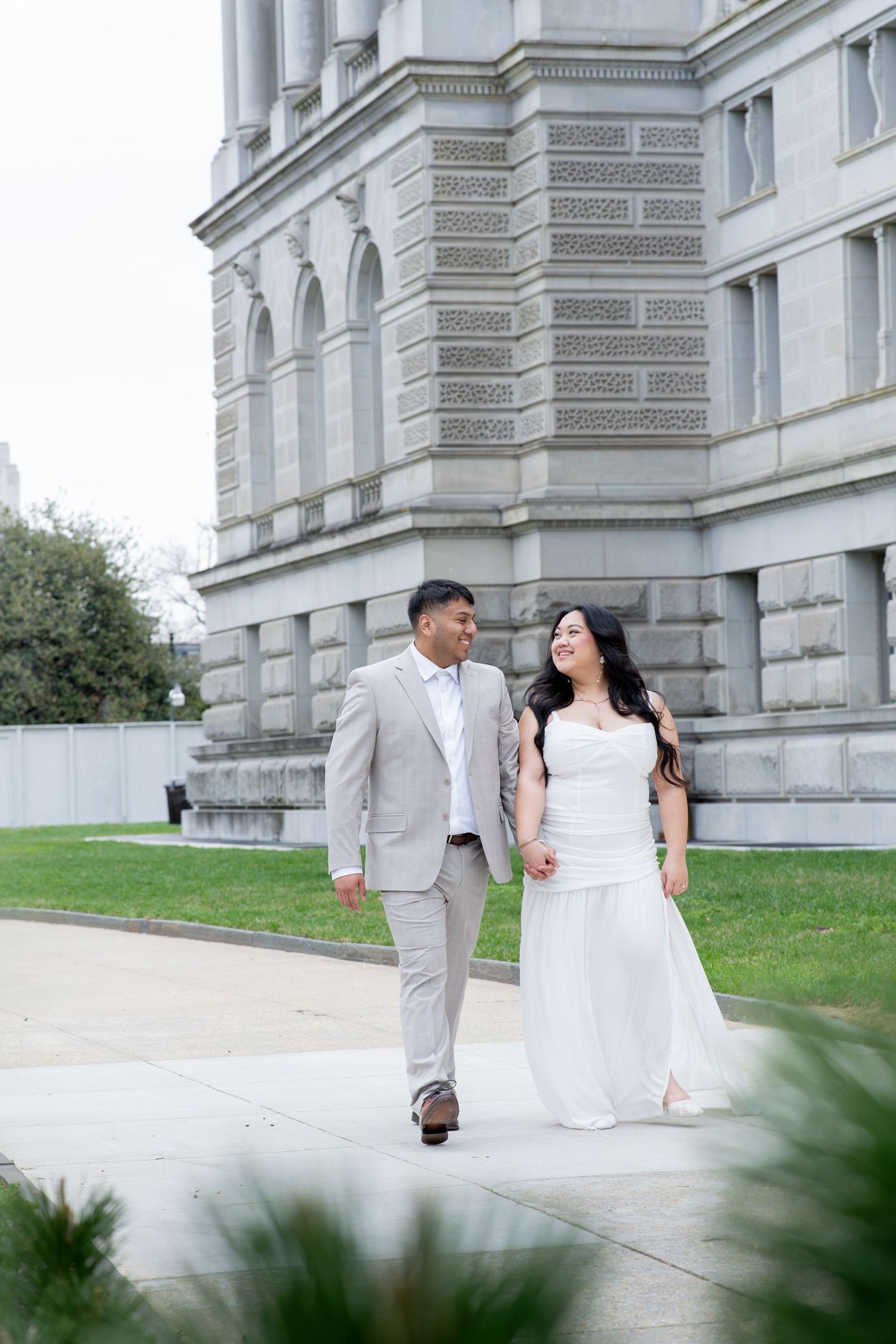 Couple in wedding attire walking hand in hand outside a grand stone building.
