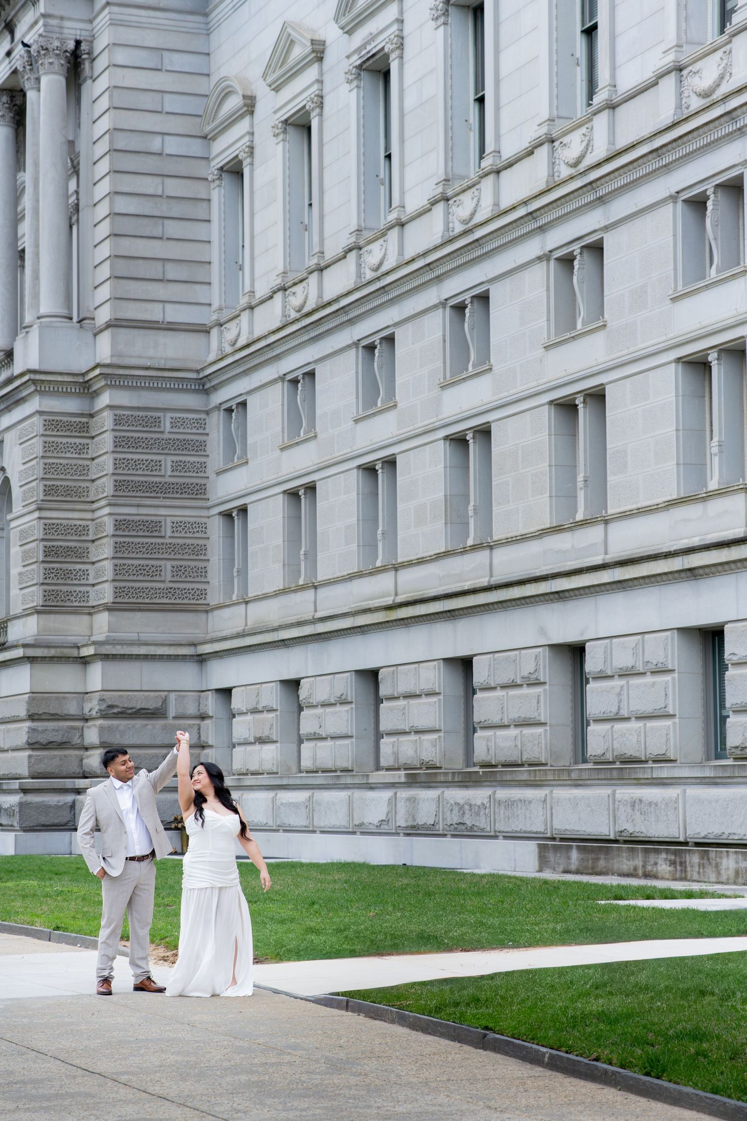 Couple in wedding attire dancing outside a grand stone government building with green lawn.