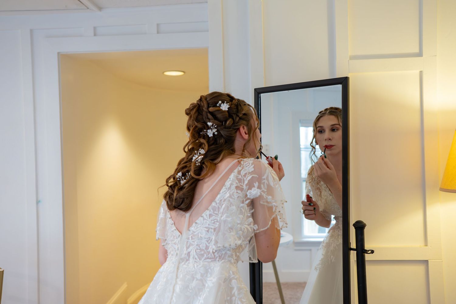 A bride in a white lace wedding dress looks at herself in a full-length mirror during the getting ready process.