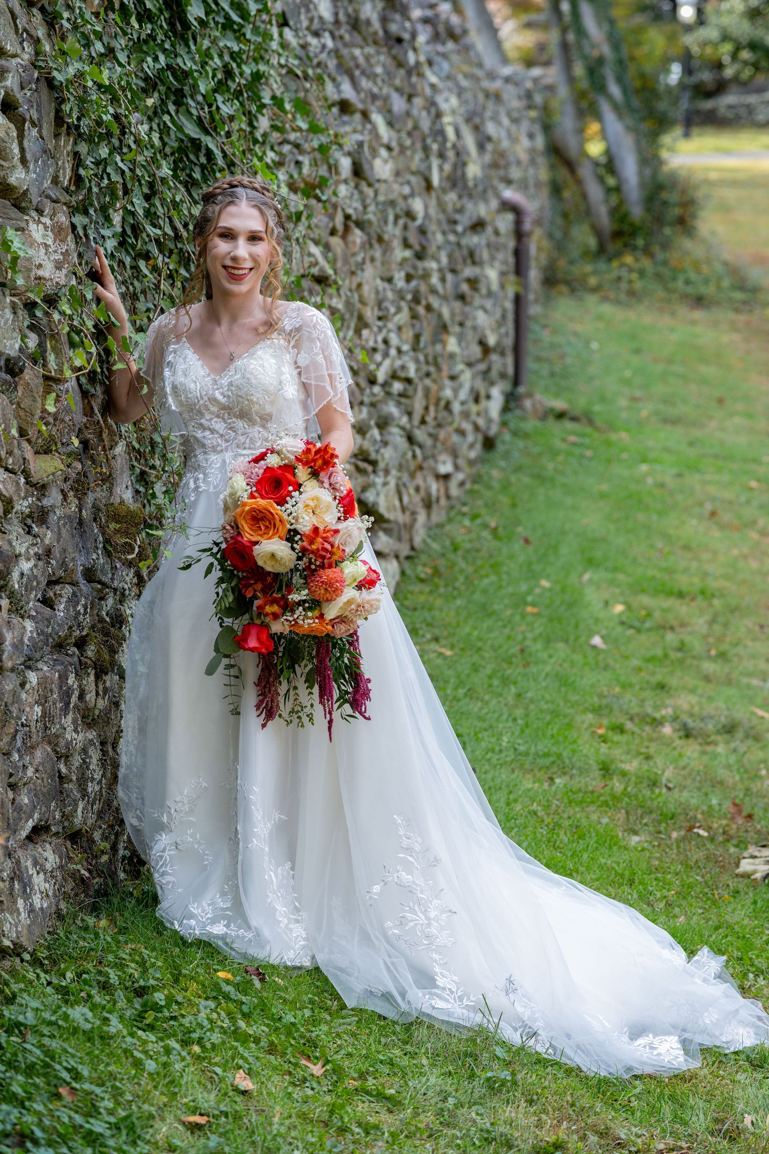 A stunning bridal portrait series against a stone wall featuring a white lace gown and vibrant red and orange autumn bouquet.