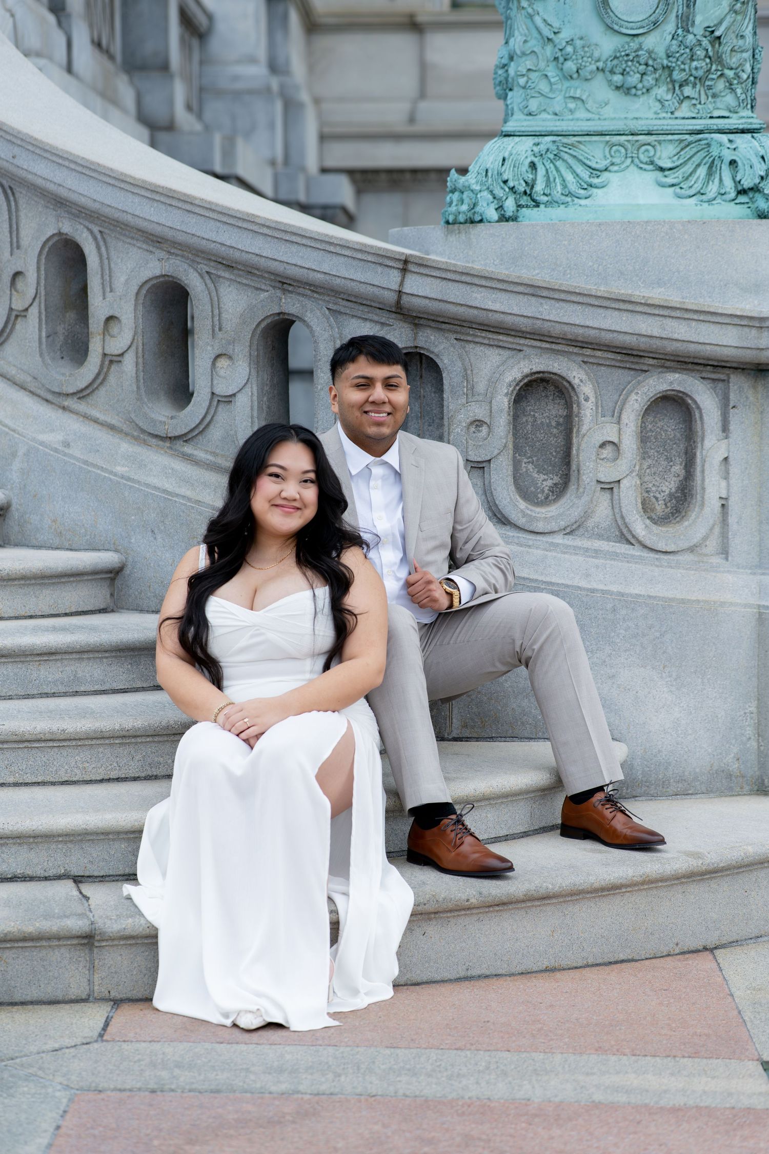 A couple in formal attire poses on ornate stone steps, woman in white gown and man in gray suit smiling.