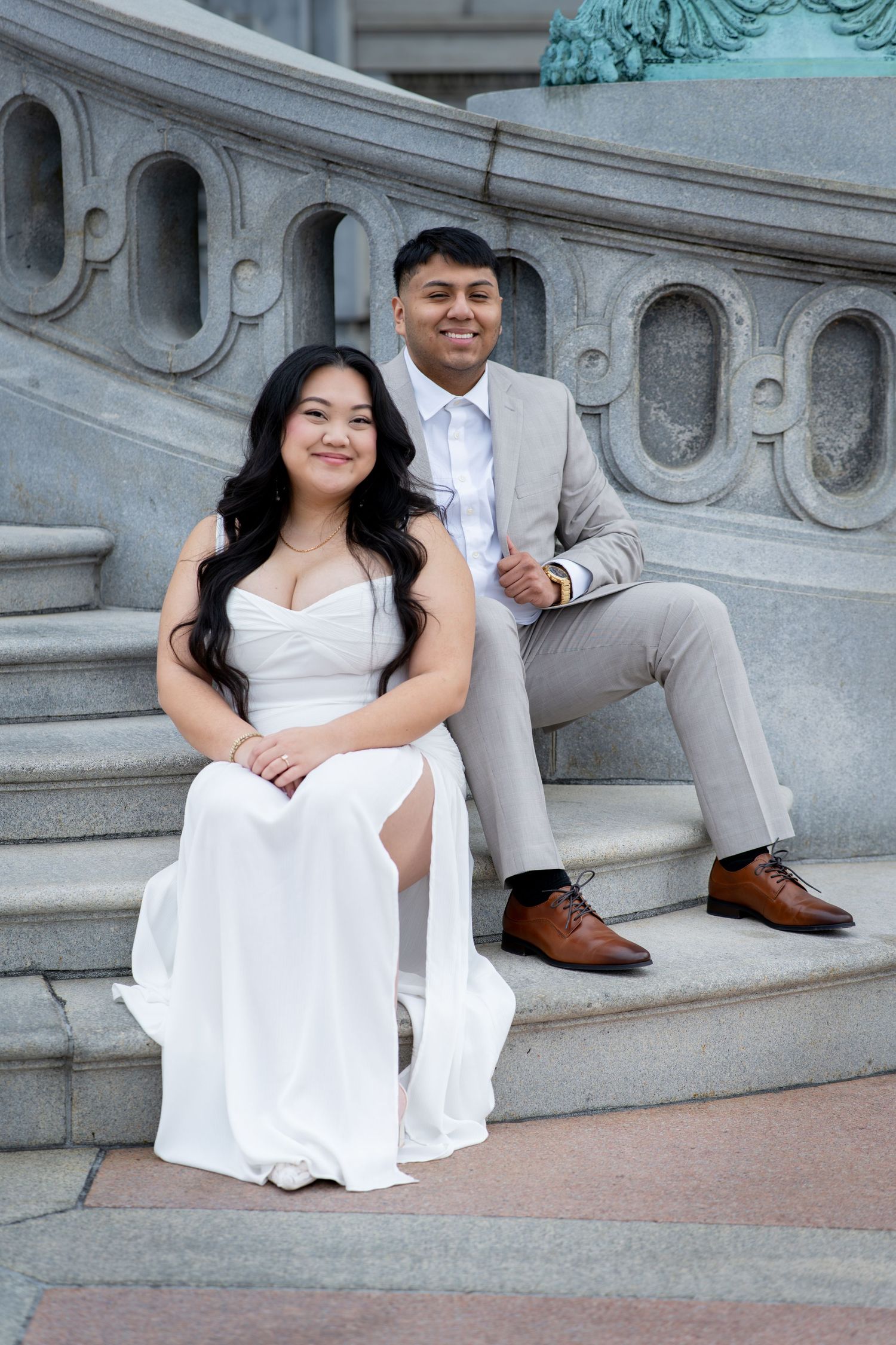 A couple in formal attire poses on ornate stone steps, woman in white gown and man in gray suit with brown shoes.
